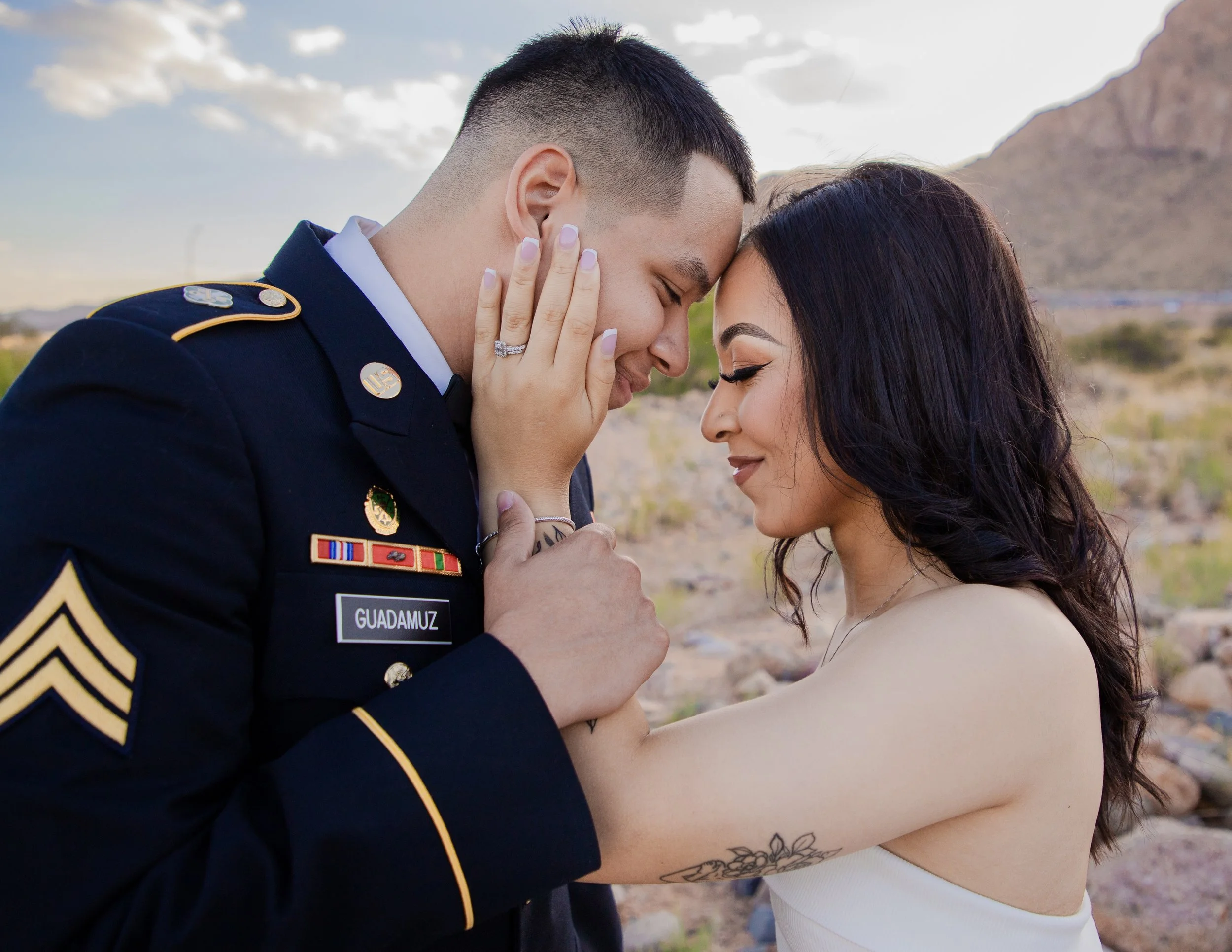 A couple shares a tender kiss outdoors, the man dressed in a U.S. Army uniform and the woman in a white sleeveless top, with a mountain landscape in the background under a partly cloudy sky.
