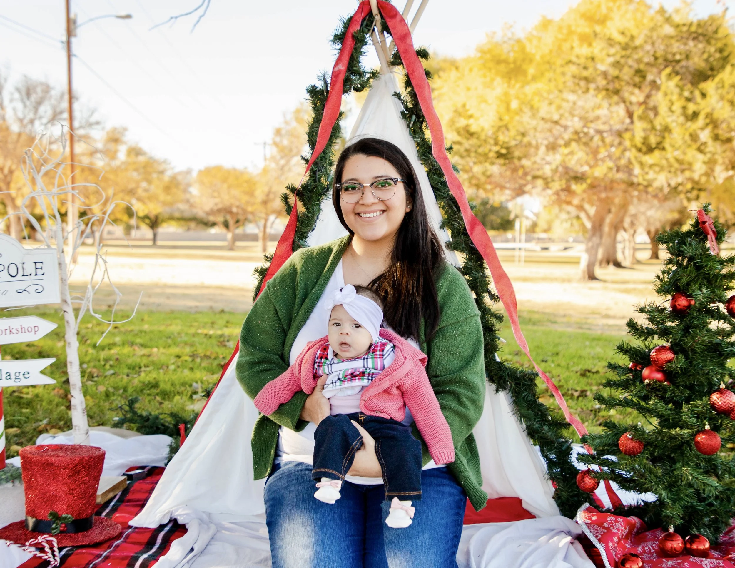 A smiling woman with glasses and long dark hair, wearing a green cardigan, holding a baby dressed in a pink sweater and plaid shirt, sitting in front of a decorated Christmas tree outdoors during autumn.