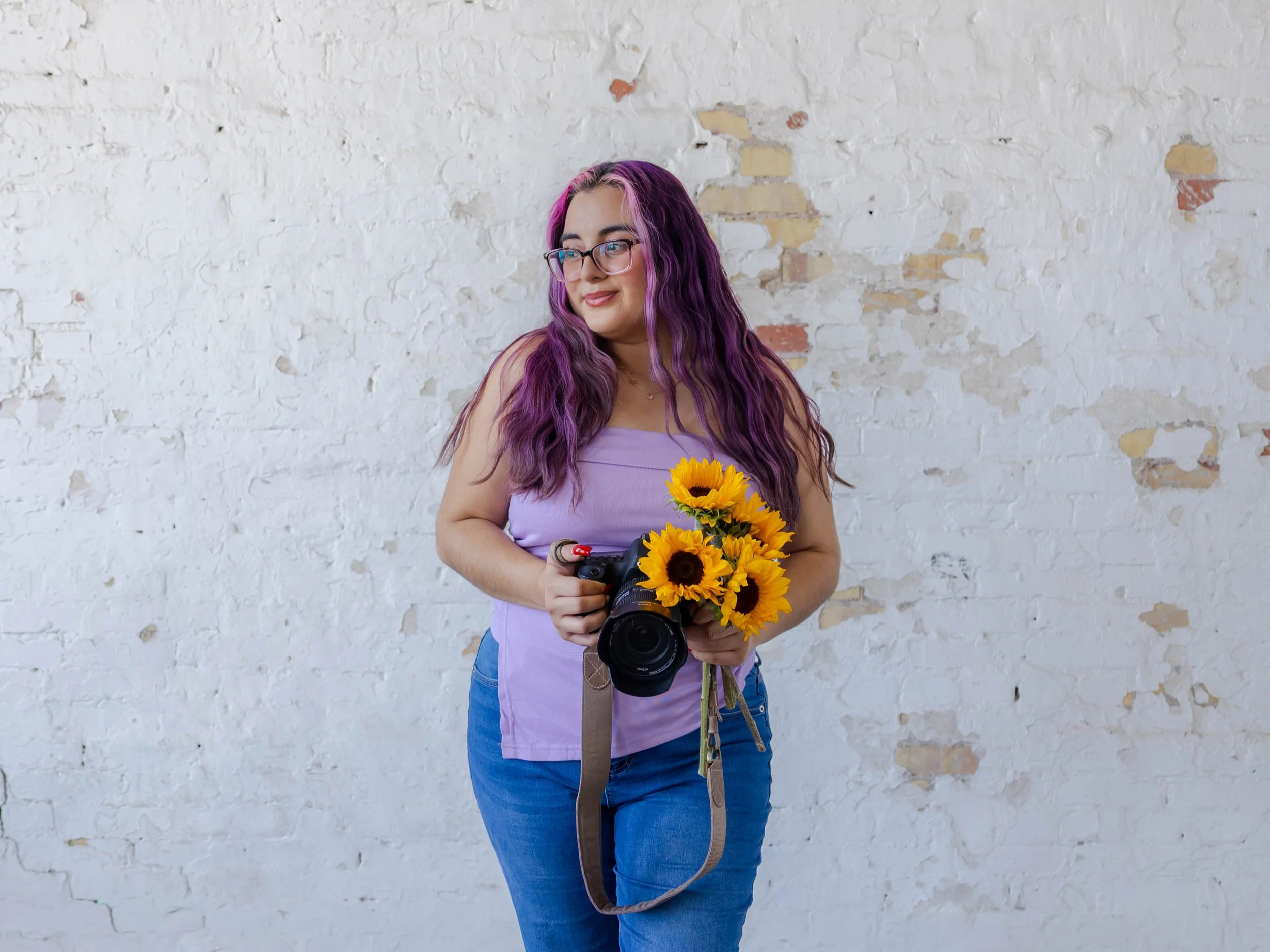 Young woman with long purple hair and glasses holding a camera and a bouquet of sunflowers standing in front of a white brick wall.