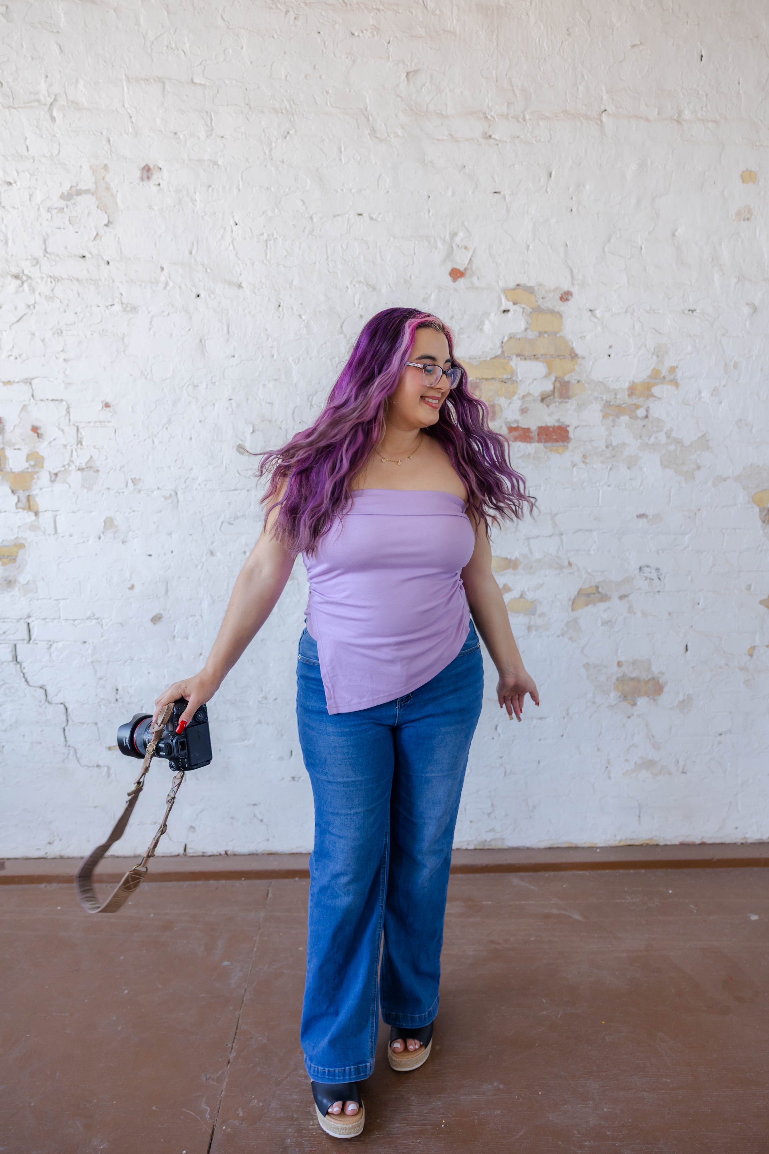 A woman with purple and pink wavy hair, wearing glasses, a lavender strapless top, blue jeans, and sandals, standing against a white brick wall with peeling paint, smiling and holding a camera with a strap in her right hand.