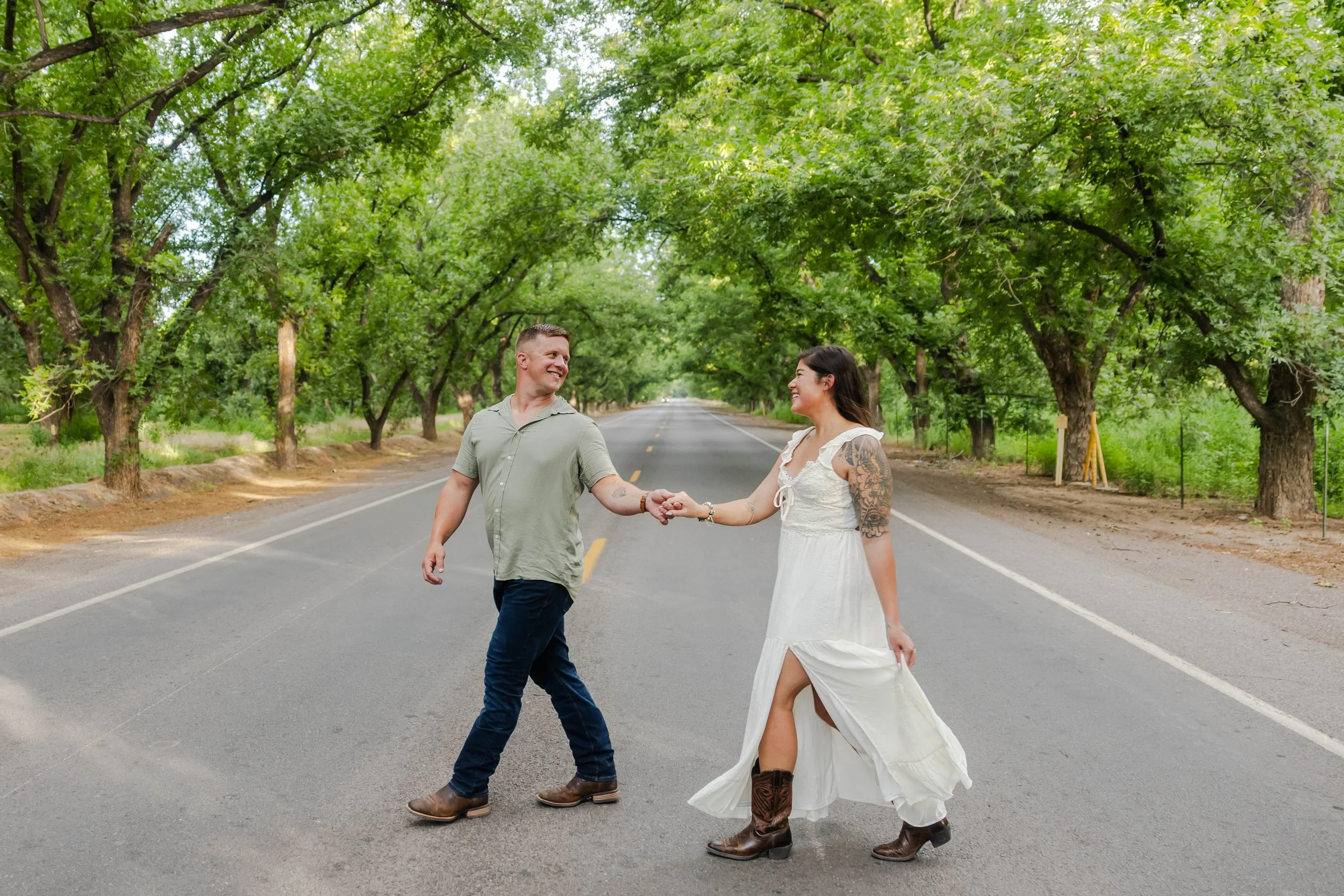 A man and woman holding hands and smiling at each other while walking across a Southern New Mexico road in the Mesilla Valley Pecan Trees.