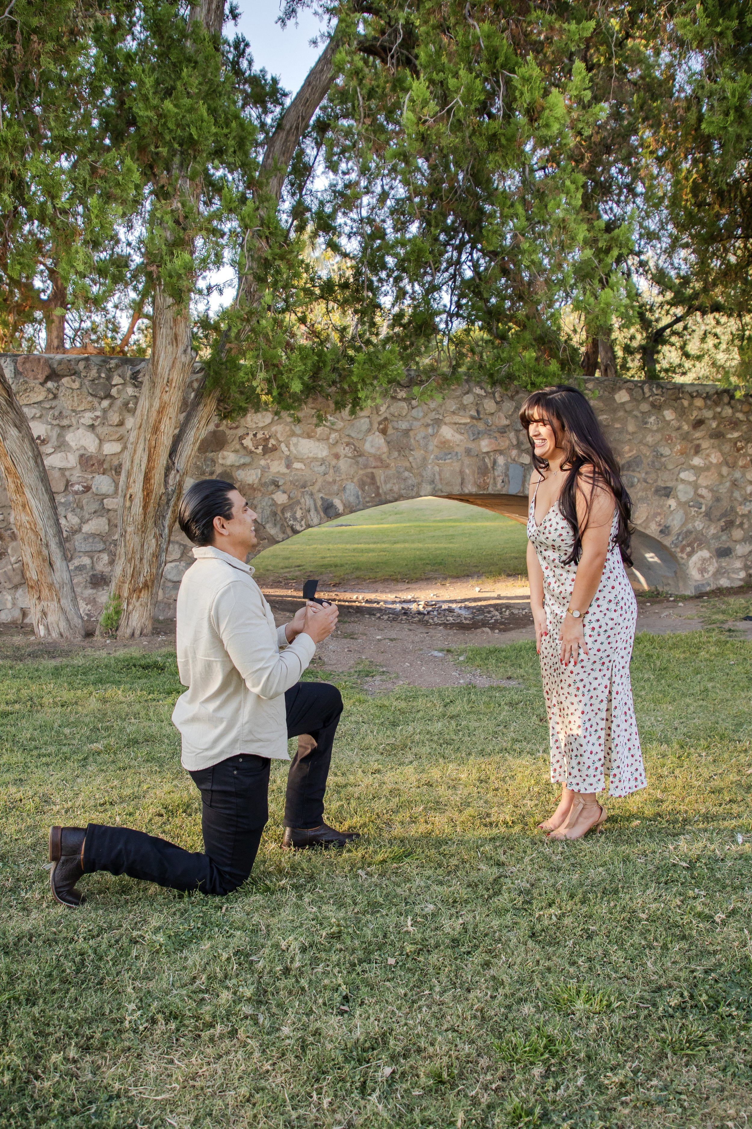 A woman with long dark hair in a white dress with red floral pattern stands smiling into the eyes of a man who is kneeling on one knee proposing with a ring in his hand, in a park with green grass, trees, and a stone bridge in the background.