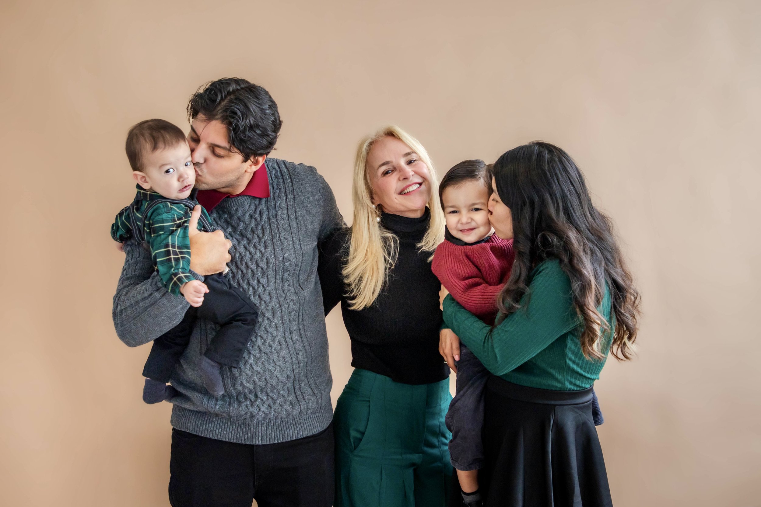 A diverse group of five people, including two men, two women, and a young boy, standing together against a beige background. One man is kissing a young boy on the cheek, and one woman is kissing a young girl on the cheek. They are smiling and appear happy.