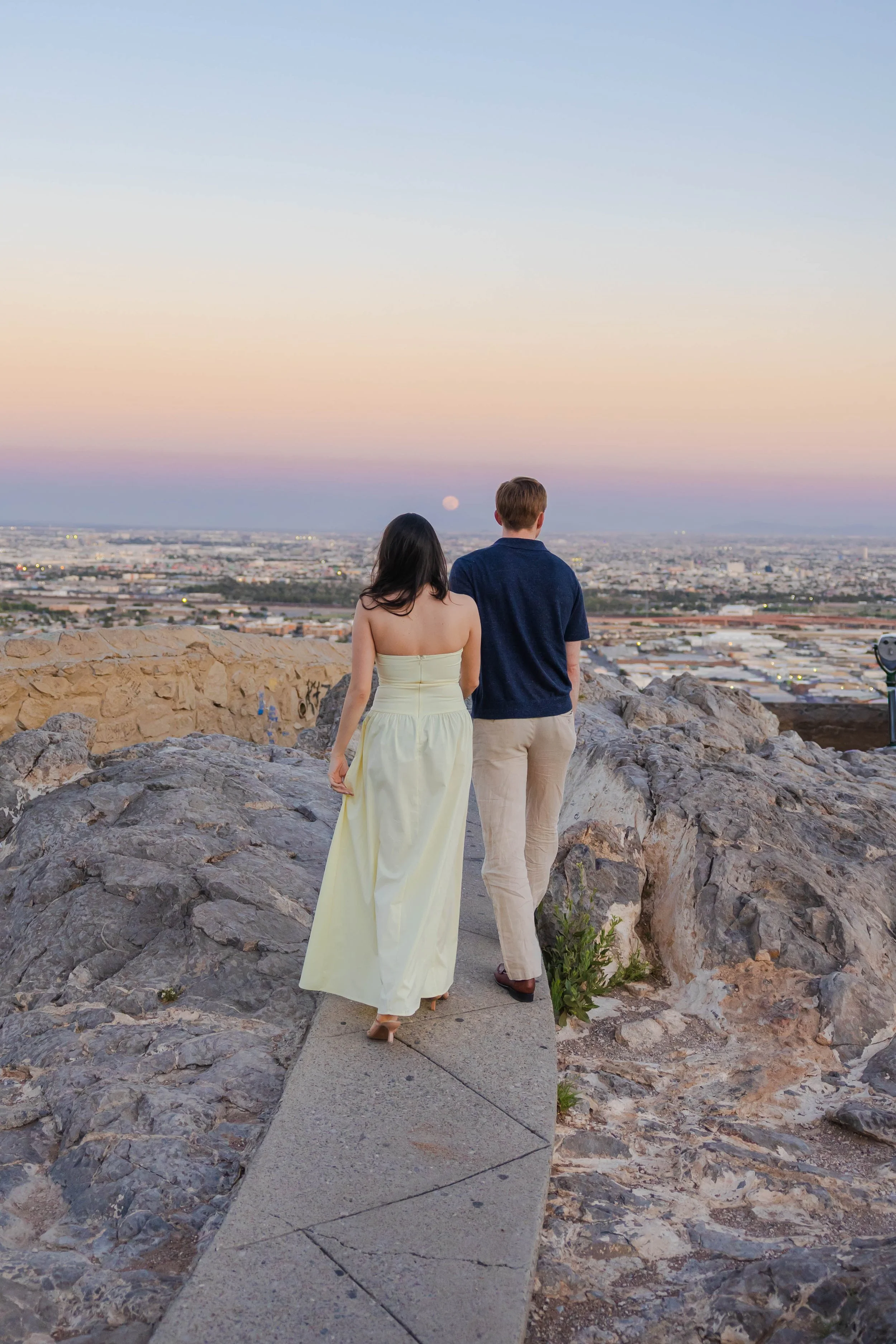 A couple stands on Scenic Drive at sunset, facing the El Paso city with the moon rising in the background, along with pastel-colored sky.