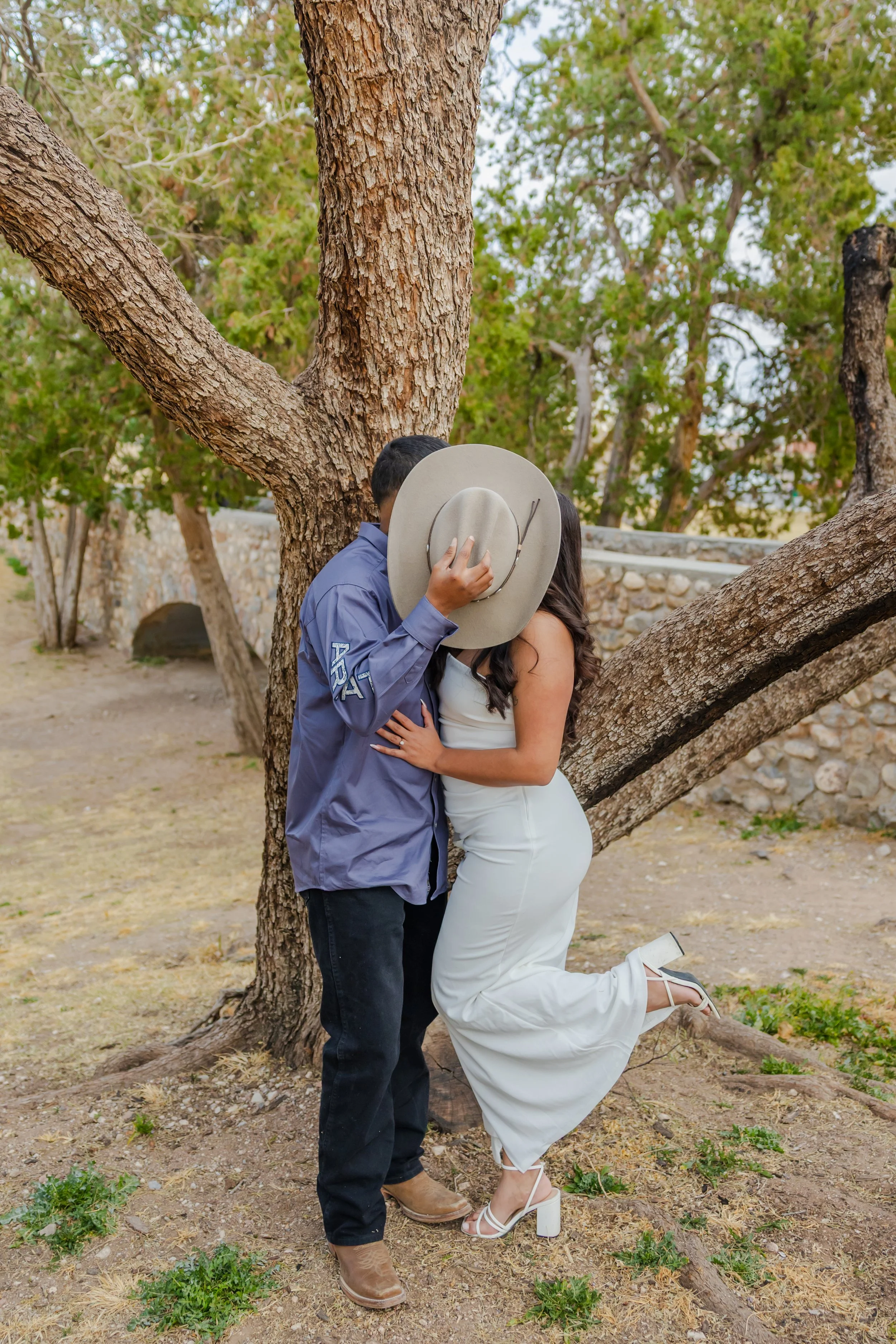 A couple is kissing under a large tree, with the man's face hidden by a wide-brimmed hat. The woman is wearing white high heels, a white dress, and has long dark hair. The man is dressed in a blue shirt with dark pants and brown boots. The scene is outdoors with a stone wall in the background.