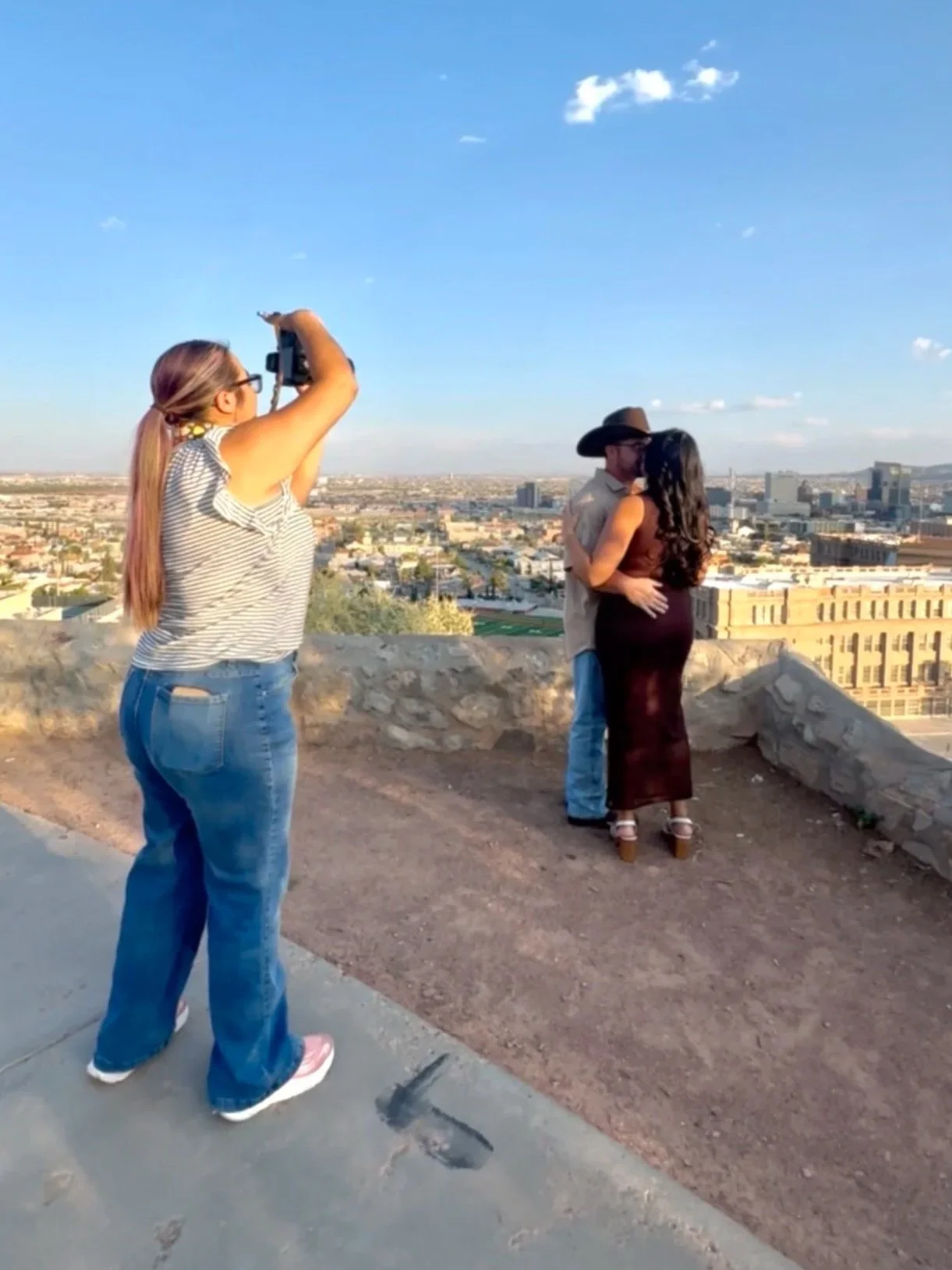 An El Paso Wedding photographer with long hair taking a photo of a man and woman kissing on a rooftop overlooking the El Paso city skyline at Tom Lea Upper Park.
