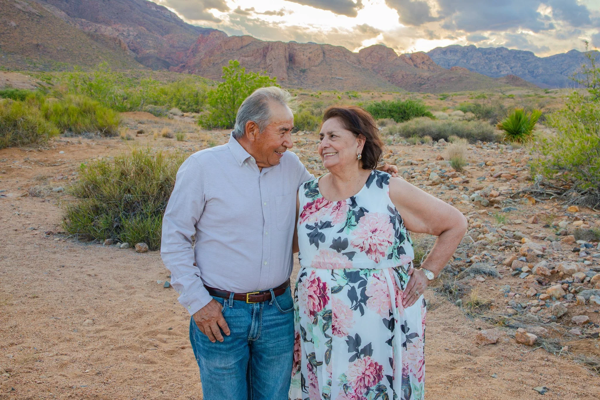 An elderly man and woman smiling and looking at each other in El Paso, Texas at the Franklin Mountains desert landscape with green shrubs and cloudy sky.