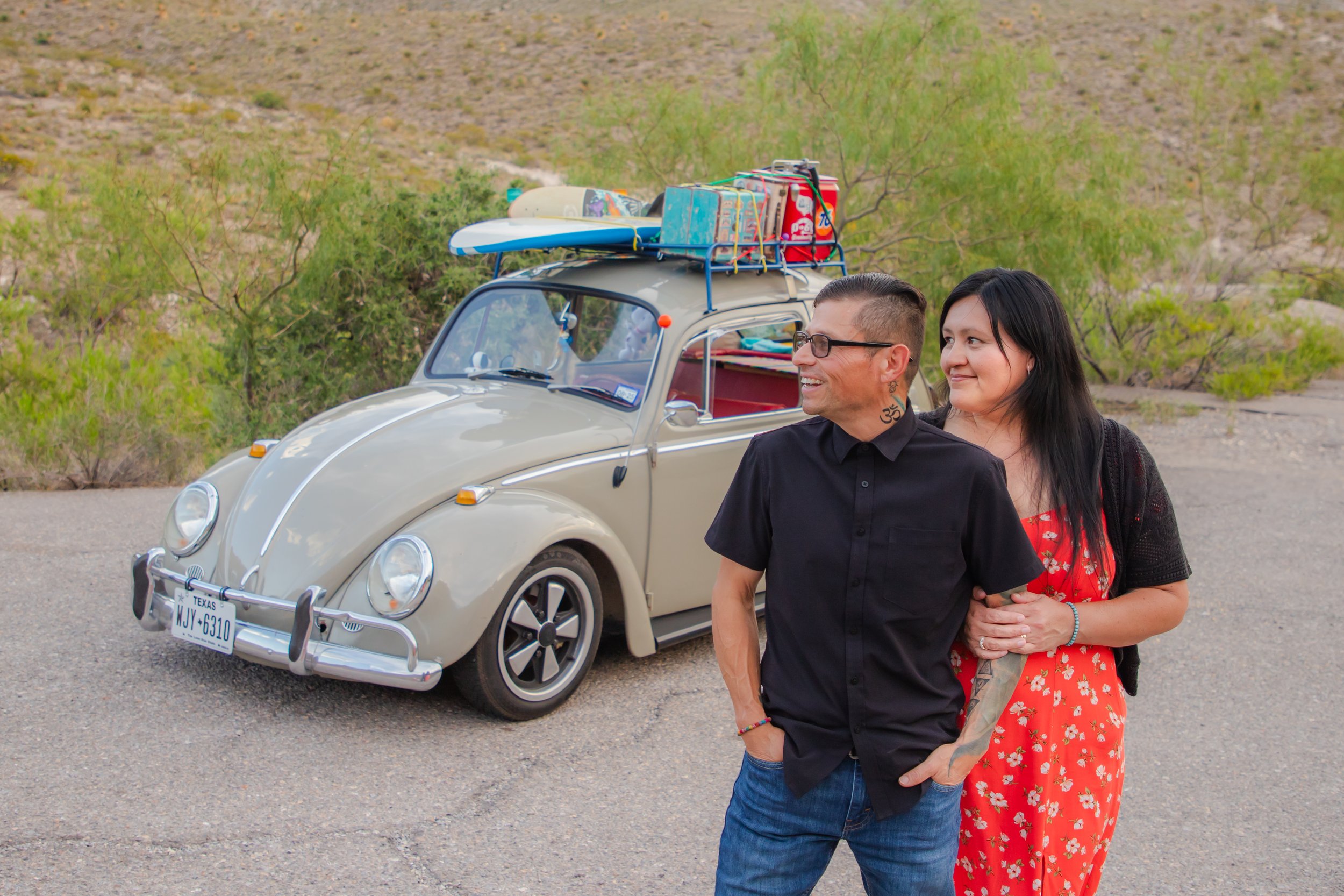A smiling man and woman walk arm-in-arm beside a vintage beige Volkswagen Beetle parked at El Paso's McKelligon Canyon with the Franklin Mountains in the background. The car has colorful luggage and a surfboard on its roof rack.