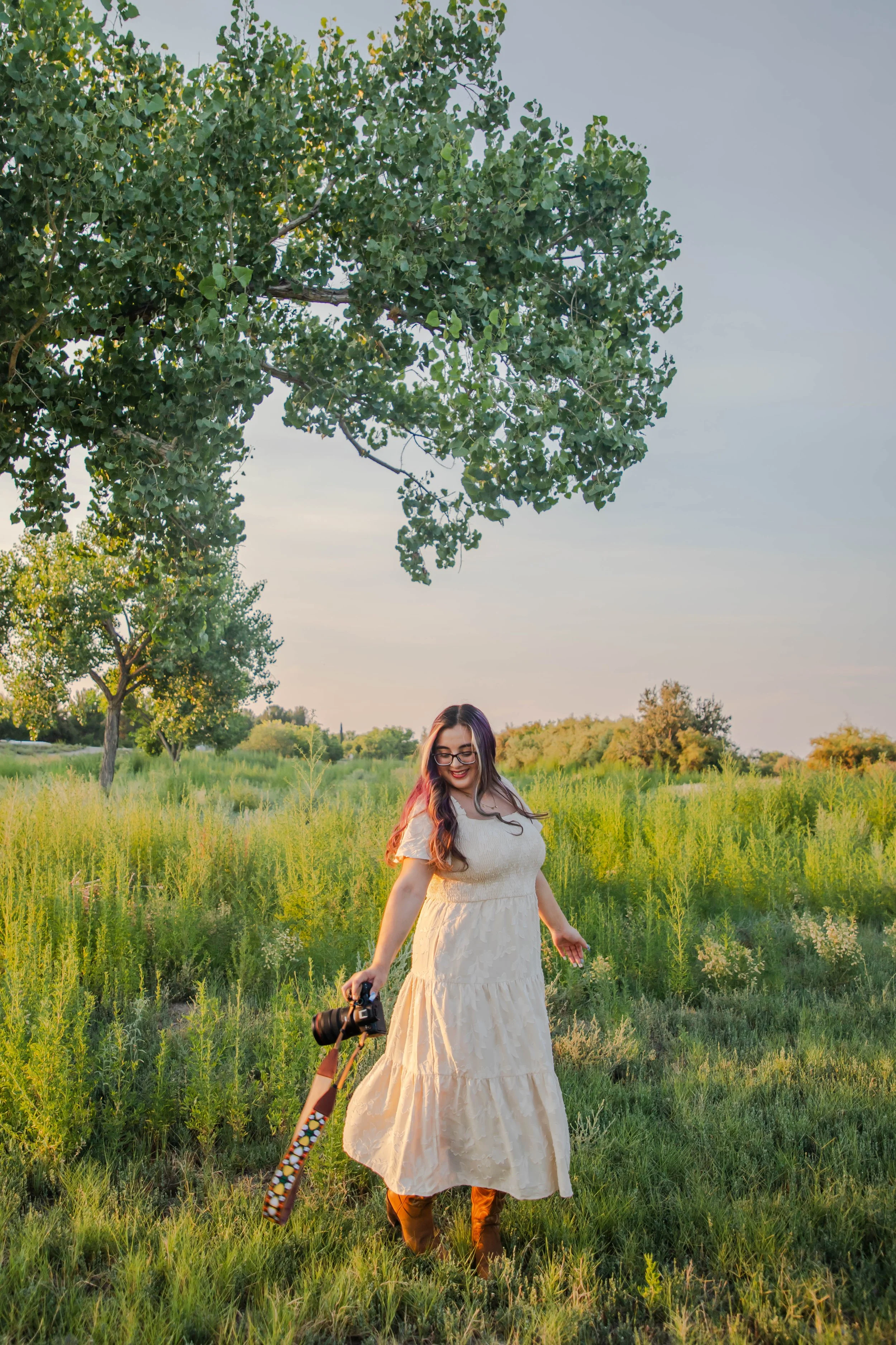 A woman with long dark hair, glasses, and a white dress walking in a green field holding a camera, with trees and a clear sky in the background during sunset.