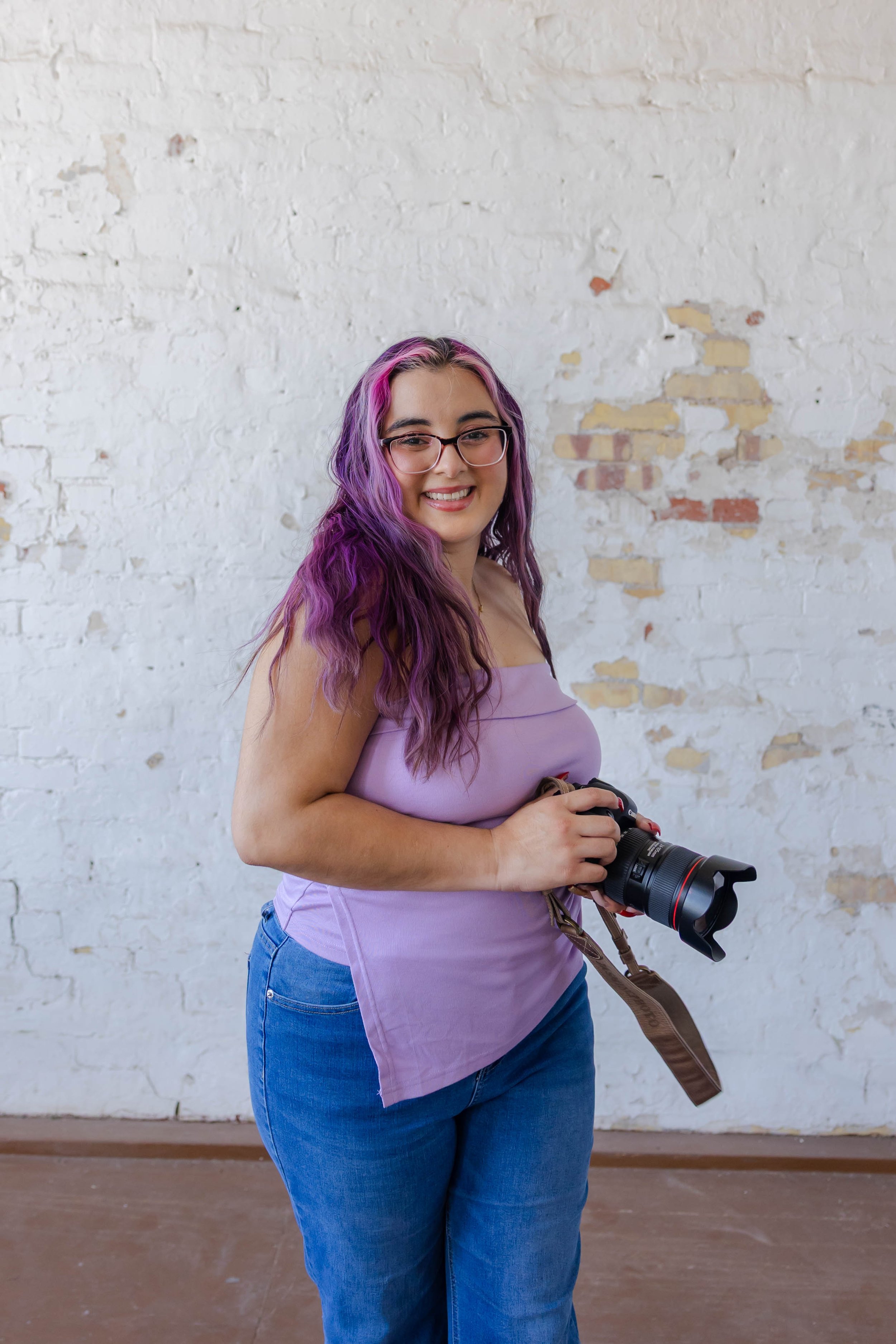A young woman with dyed pink and purple hair, wearing glasses, a lavender top, and jeans, holding a professional camera, standing in front of a white brick wall.