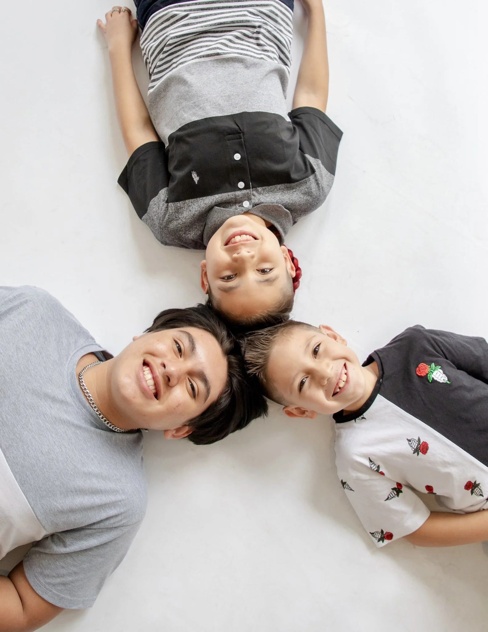 A young boy lying down on a white floor, smiling and looking up at the camera. He is wearing a gray and black shirt and patterned shorts. Two women are partially visible next to him, also lying down, with their heads near his, smiling.