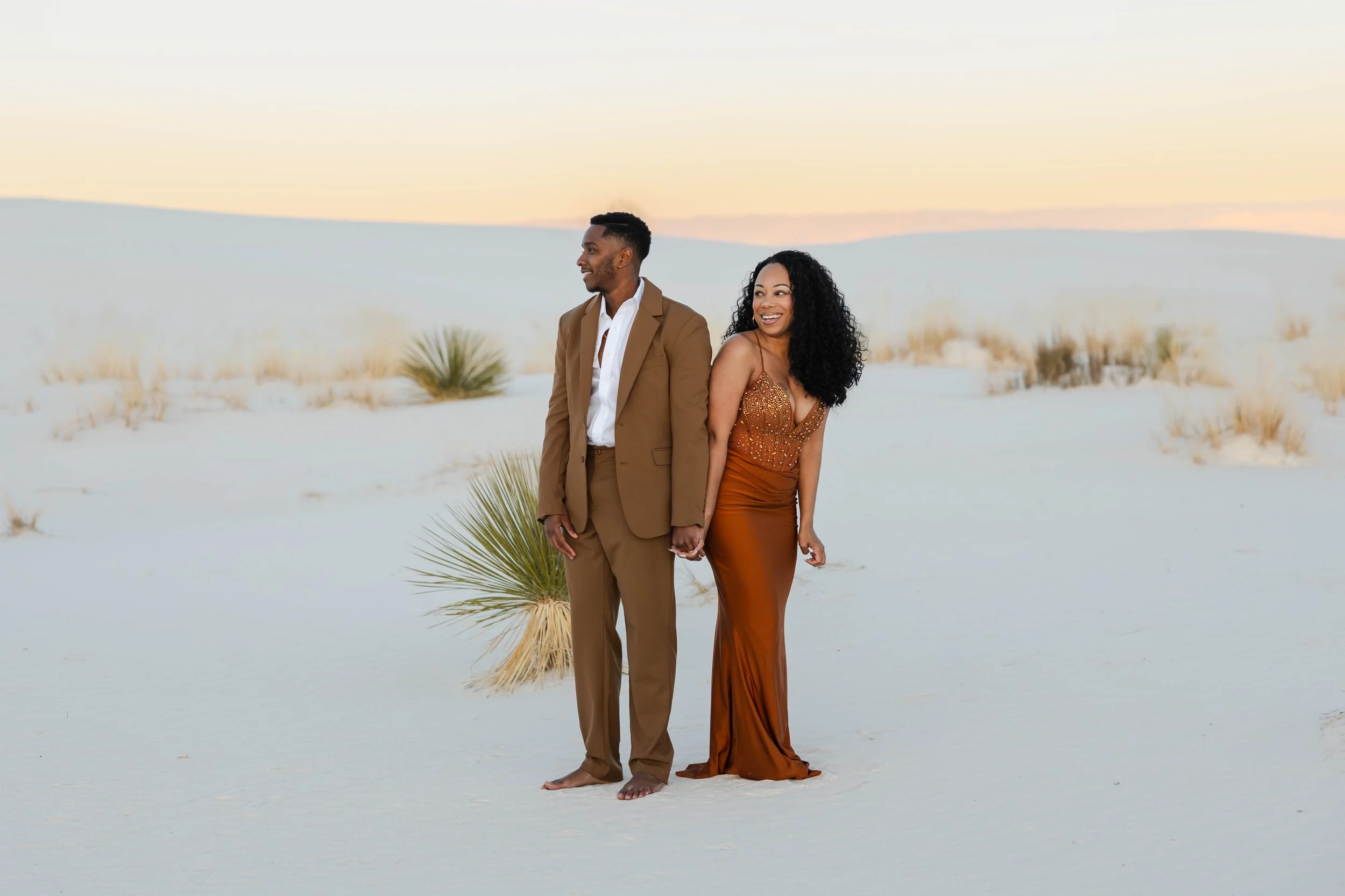 A man and woman hold hands and smile while standing barefoot on a sandy desert landscape during sunset. The man is dressed in a brown suit, and the woman is in a bronze-colored, glittery dress.