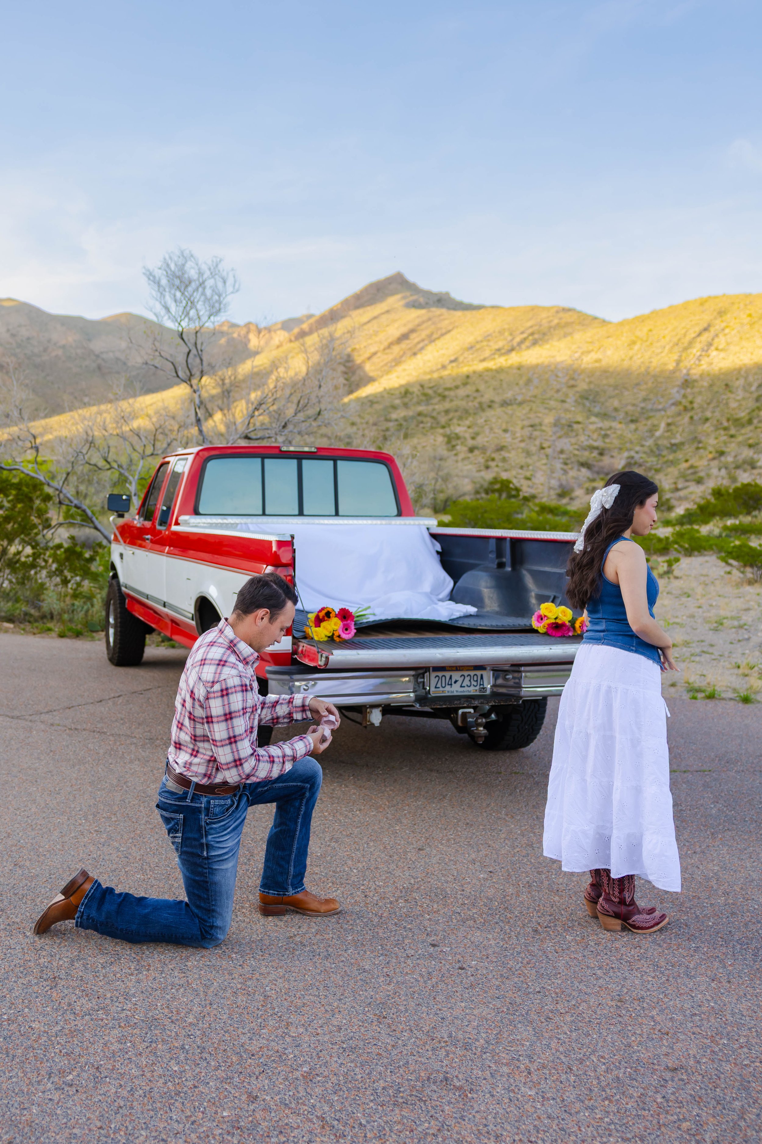 A man kneeling and praying in front of a woman in a white skirt and dark blue top, with a man kneeling next to him in front of a decorated pickup truck in a mountainous landscape.
