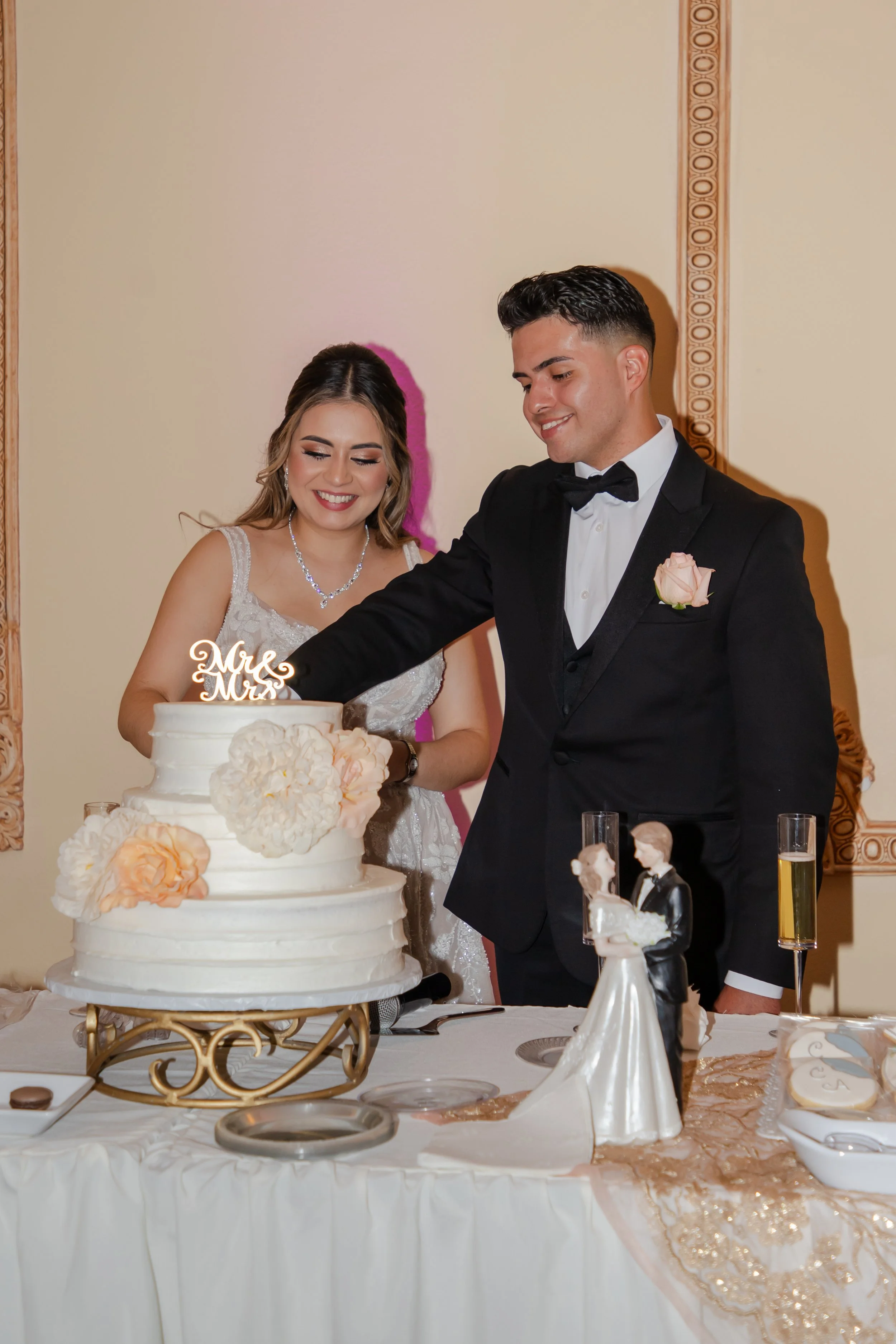 A bride and groom are laughing while cutting a wedding cake at a reception. The bride is wearing a lace wedding dress and jewelry, and the groom is in a black tuxedo with a bow tie and a pink flower boutonniere.