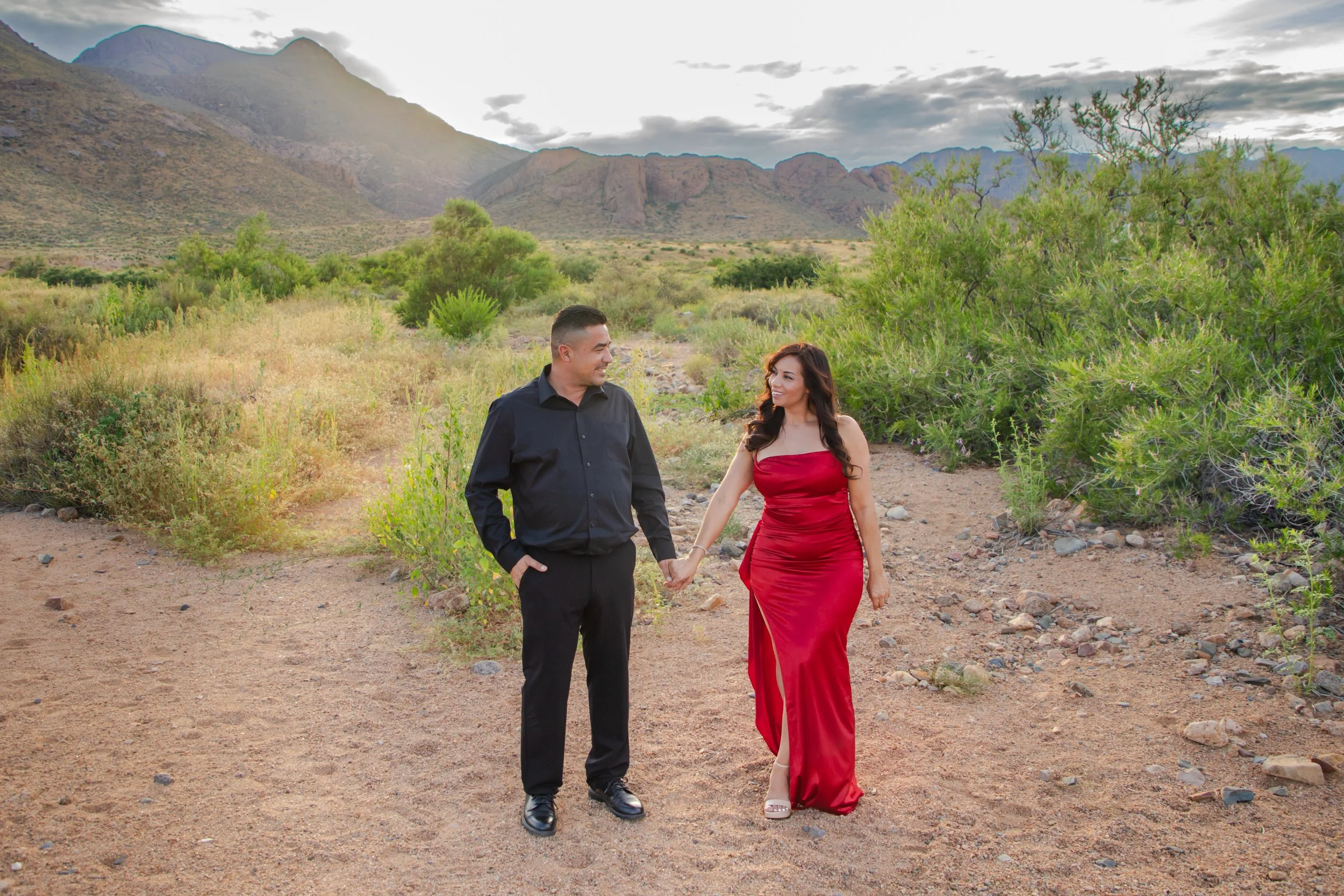 A man and woman holding hands and walking outdoors in the Franklin Mountains desert landscape near the El Paso Museum of Archaeology. The man is dressed in a black shirt and pants, while the woman wears a red satin evening gown.
