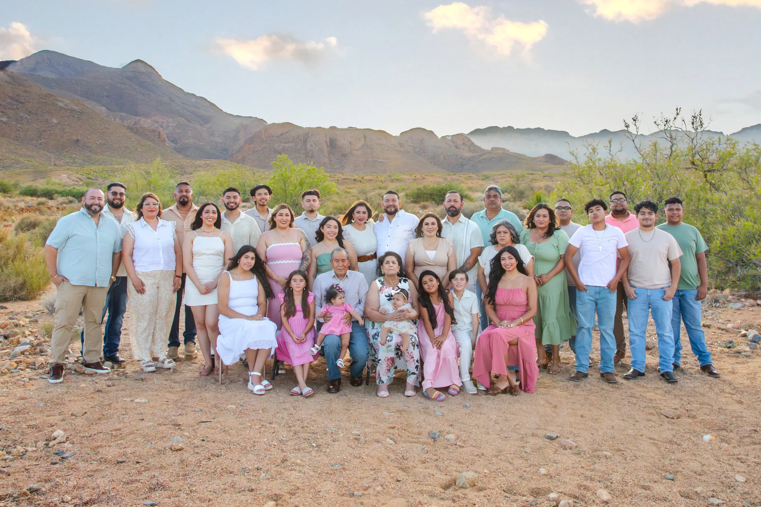 A large group of people posing for a photo outdoors with a mountainous landscape in the background. The group includes men, women, and children dressed in casual and semi-formal clothing, standing and sitting on rocky ground.