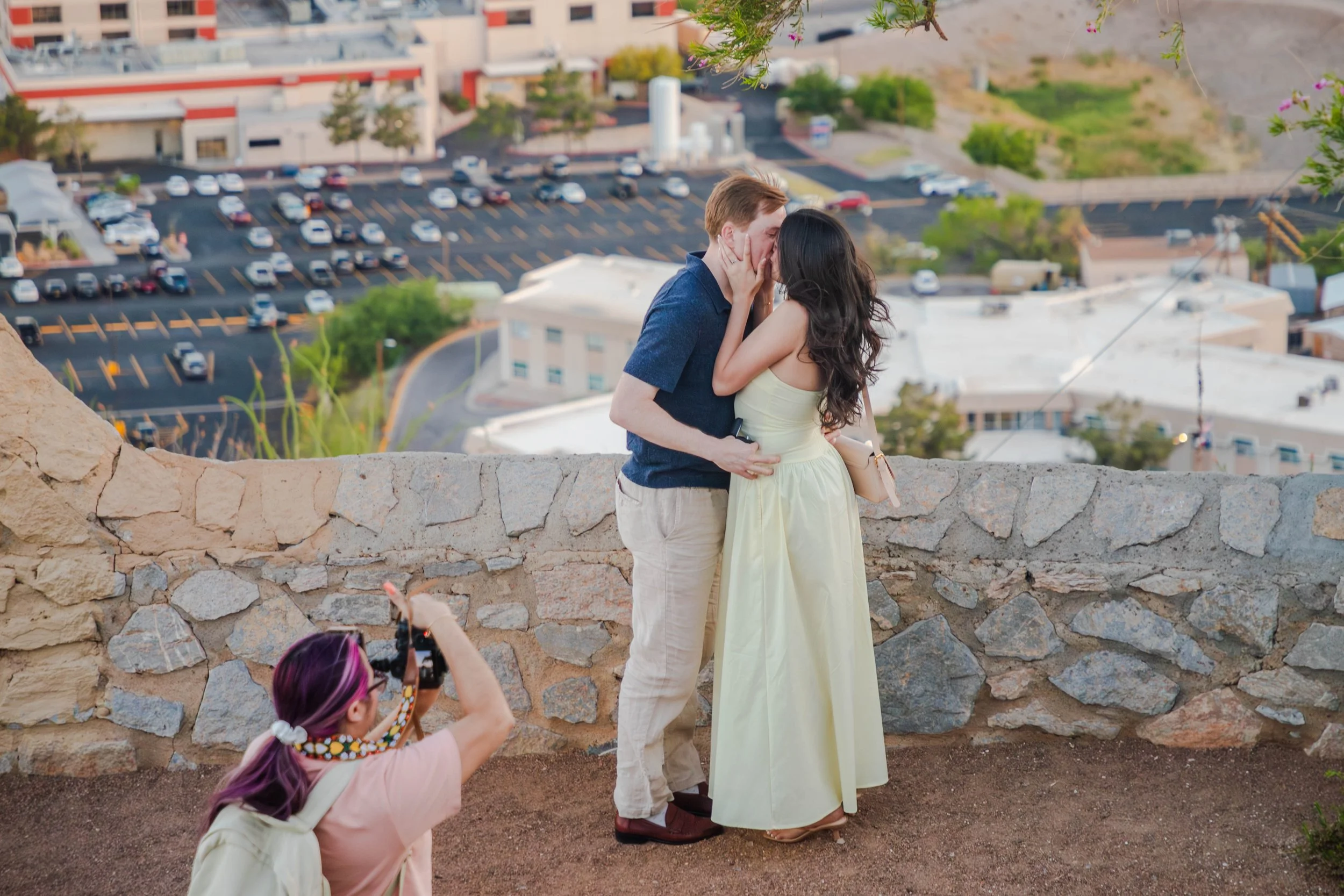 An engaged couple is kissing on a stone wall at the El Paso scenic drive overlook. A girl with purple hair and a flower crown is taking a photo of them.