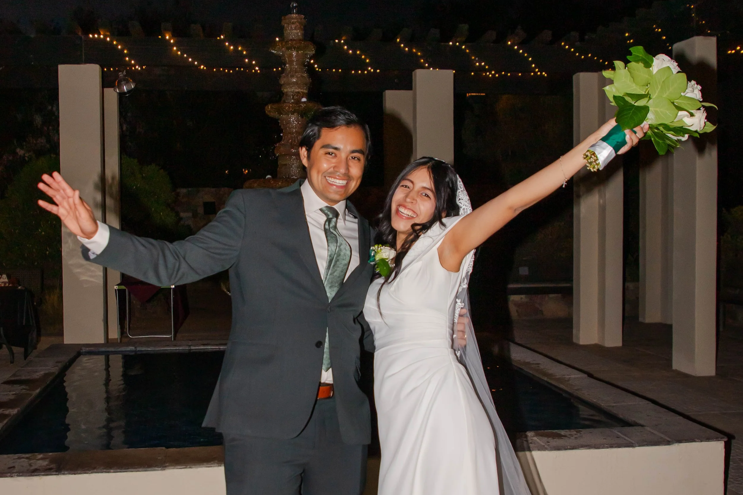 A couple dressed in wedding attire, smiling and celebrating at night near a water fountain at Keystone Heritage Park, with string lights overhead, holding a bouquet of green and white flowers.