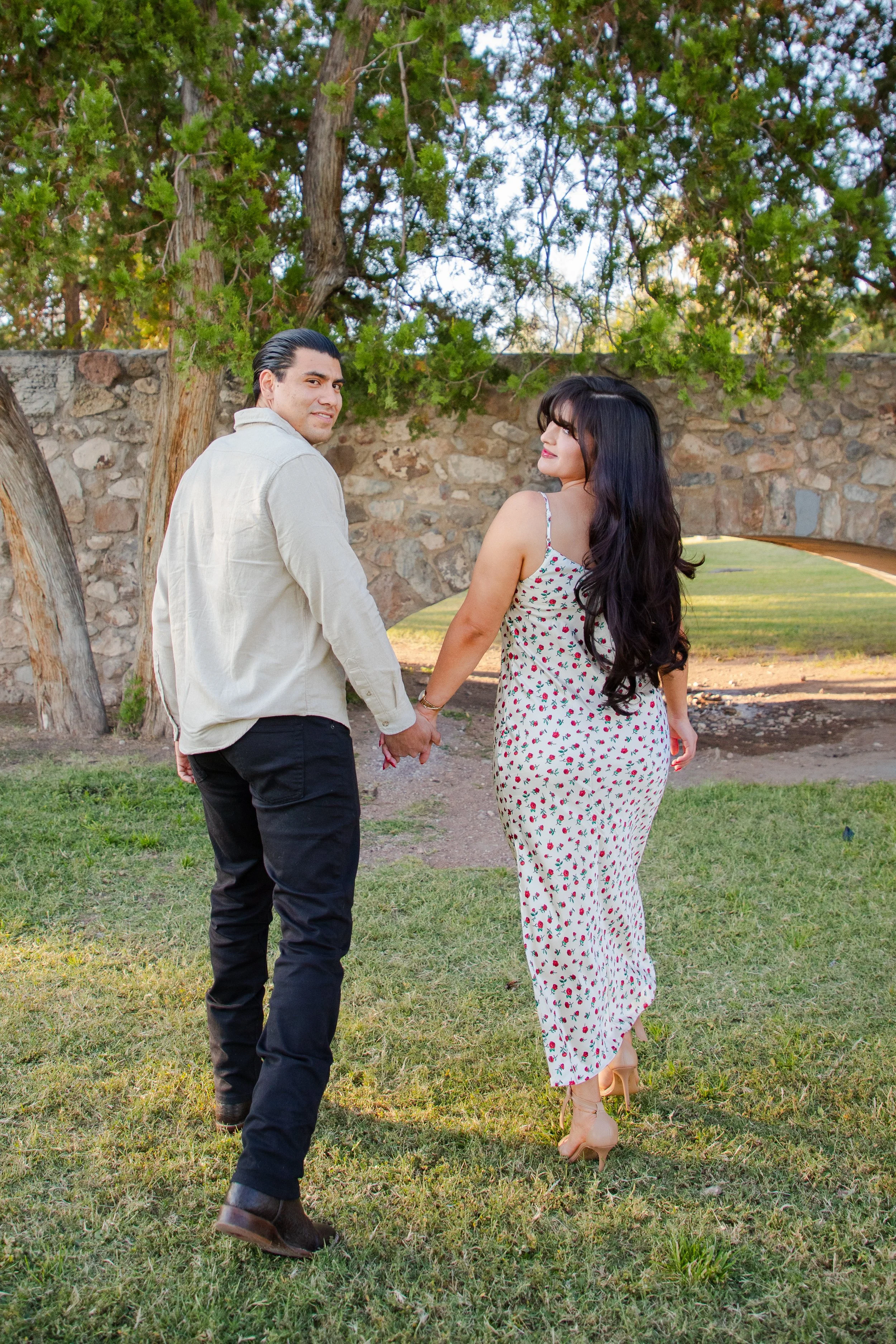 A couple holding hands and walking in El Paso's Memorial Park on a grassy area with trees and a stone wall in the background.