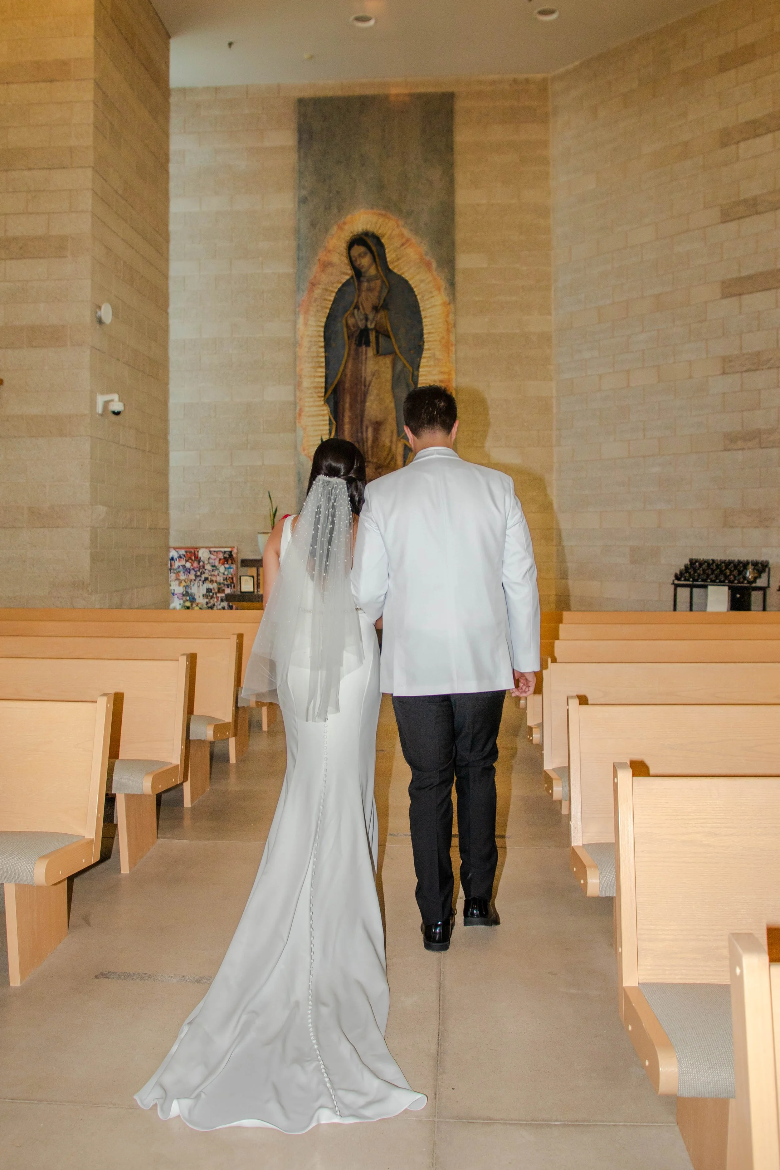 A bride and groom walk down the aisle at El Paso's St. Stephens Catholic Church, with the bride wearing a white wedding gown with a long train and veil, and the groom in a white jacket, heading towards a painting of the Virgin Mary on the wall.