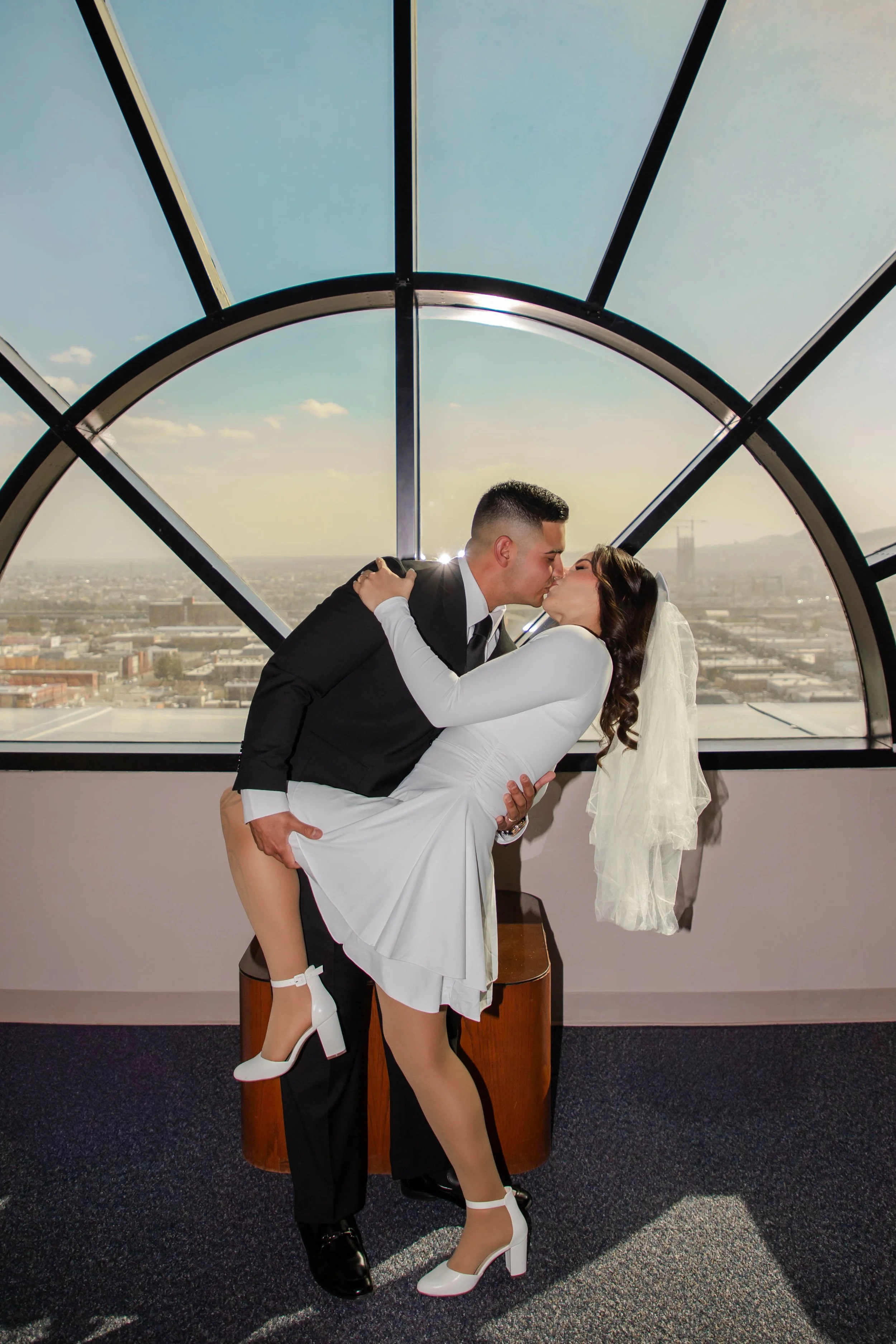 A bride and groom sharing a kiss inside the Enrique Moreno Courthouse with the El Paso city view in the background. The groom is carrying the bride, who is wearing white high heels and a white dress.