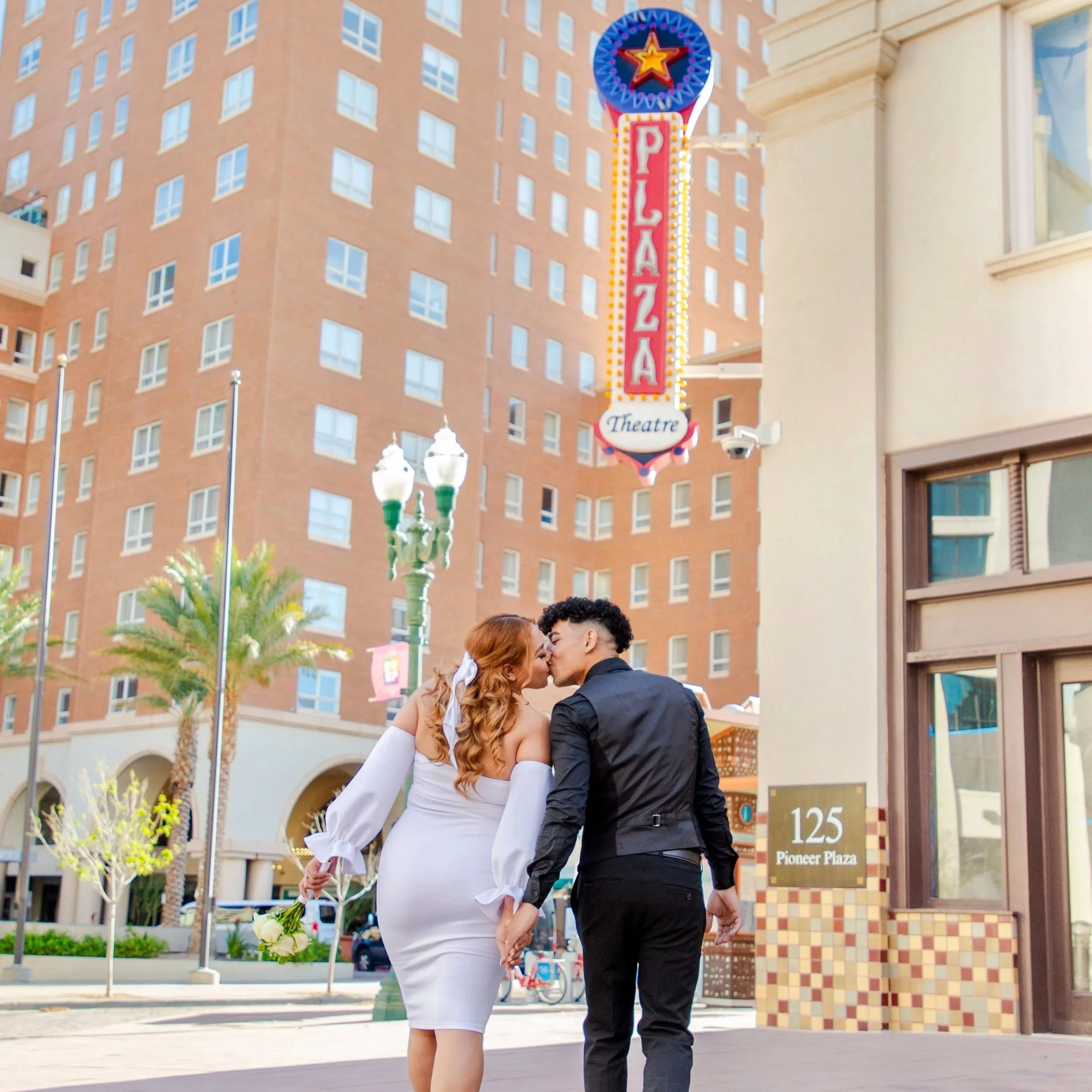 A couple kissing during their wedding, holding hands, with a bouquet of flowers in the woman's hand, standing outdoors in front of El Paso's Hotel Paso del Norte building and street signs.