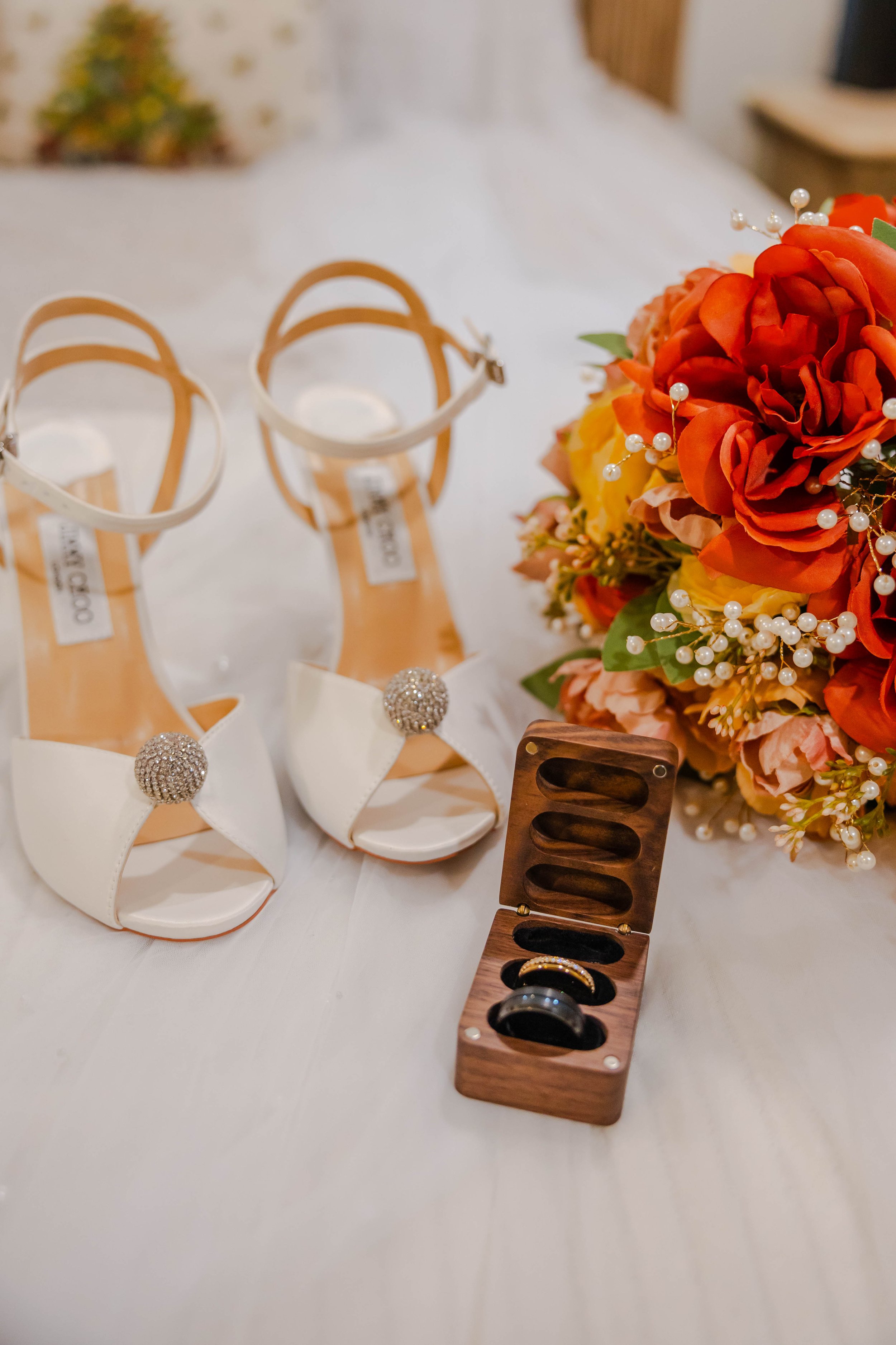 Pair of white high-heeled shoes with rhinestone decoration, a wooden ring box containing wedding bands, and a bouquet of orange and yellow flowers with pearl accents on a table.