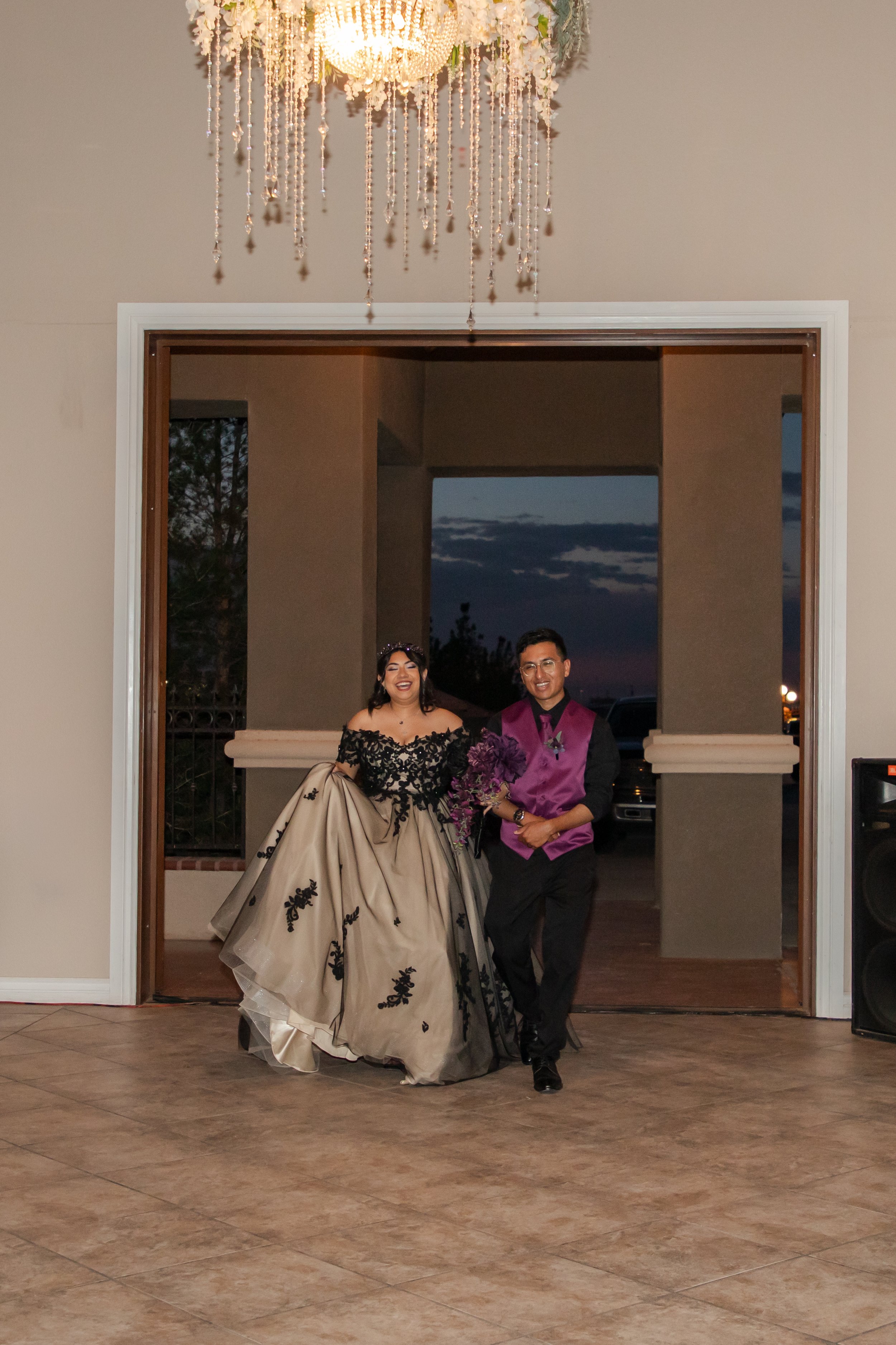 An El Paso couple just married near a large open doorway. The woman is wearing a strapless black and beige ball gown with floral embroidery, and the man is in a dark suit with a purple vest and shirt.