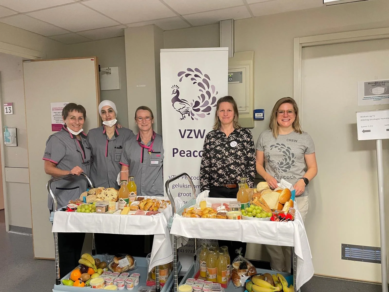 Vijf mensen staan achter een tafel met ontbijtproducten. Ze lachen en dragen verschillende kleding: drie in medische uniformen, twee in casual kleding. Op de achtergrond staat een bord met de tekst 'VZW Peace'. Op de tafel liggen brood, fruit, sap, e