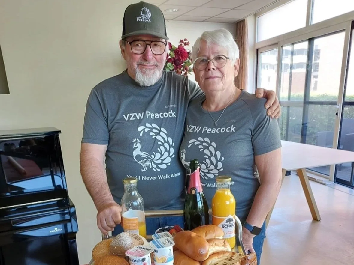 Een oudere man en vrouw staan samen achter een tafel met ontbijt. Ze dragen grijze t-shirts met 'VZW Peacock' en de slogan 'You'll Never Walk Alone'. Ze glimlachen, en er staan ontbijtproducten zoals brood, fruit, sap en een fles champagne op de tafe