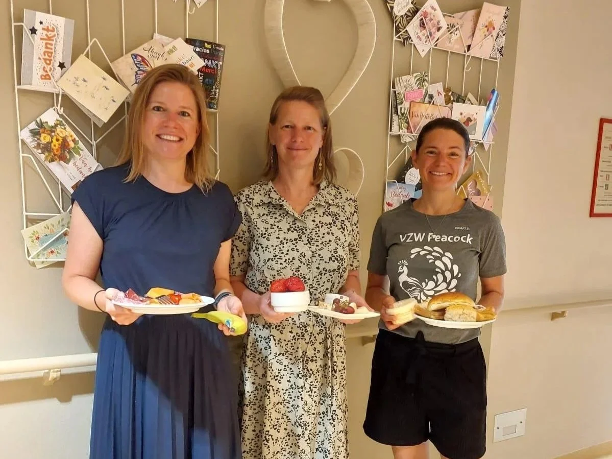 Drie vrouwen staan samen, lachen en houden schalen met desserts en sandwiches. Achter hen een muur met kaarten en een hartvormig decor.