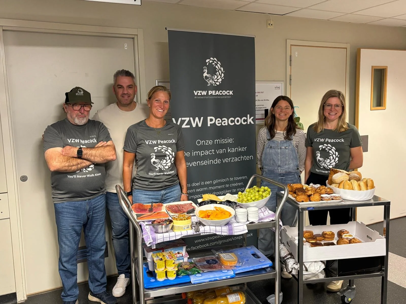Vijf mensen staan achter een tafel met eten, waaronder brood, fruit, vlees, en gebak. Ze dragen T-shirts met de tekst 'VZW Peacock' en een pauw logo. Er staat een banner met dezelfde tekst en een missieomschrijving.