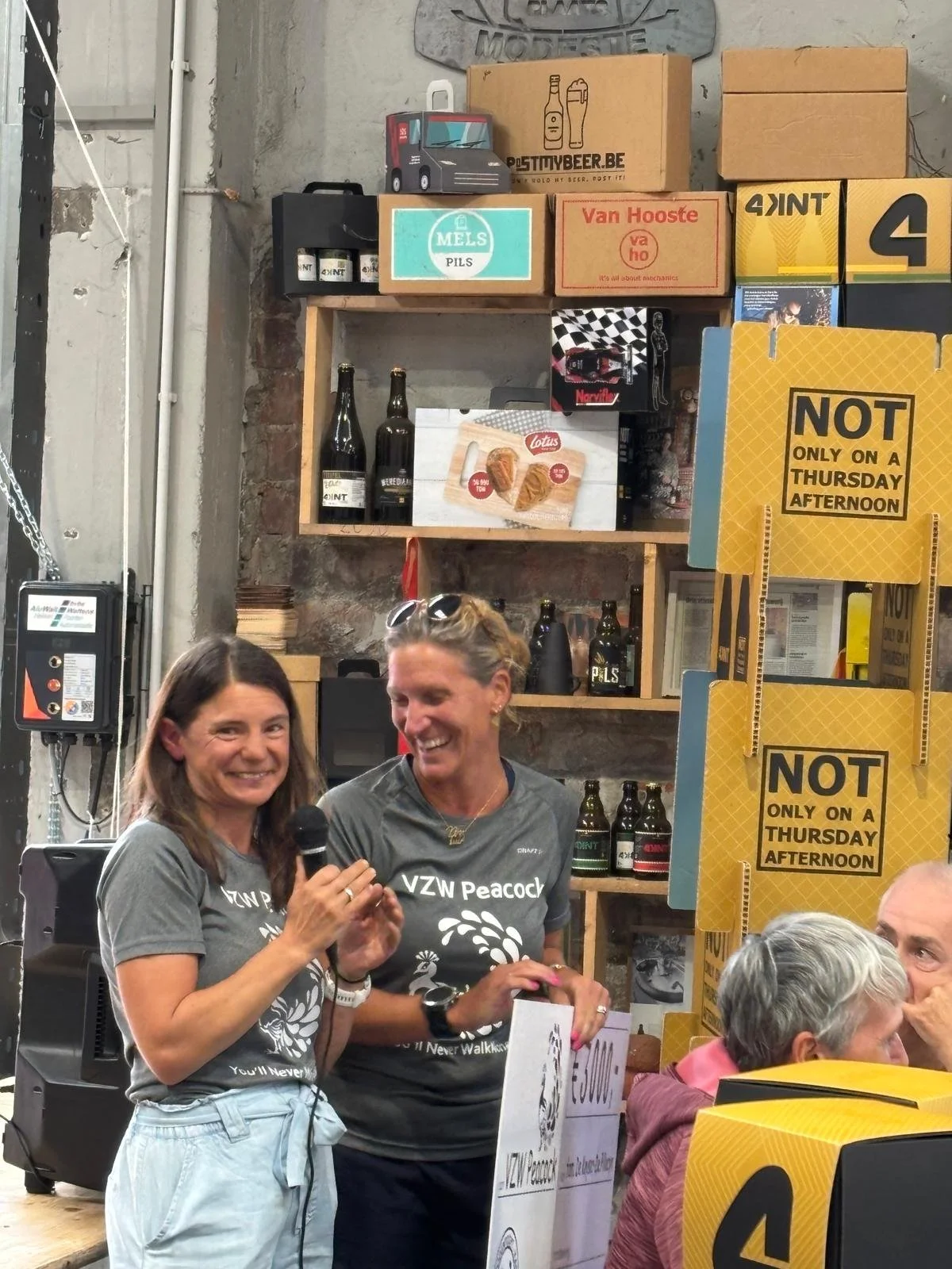 Twee vrouwen houden een microfoon en lachen, achter hen een muur met biervaten en voorraadkasten, en een groep mensen zit aan een tafel.