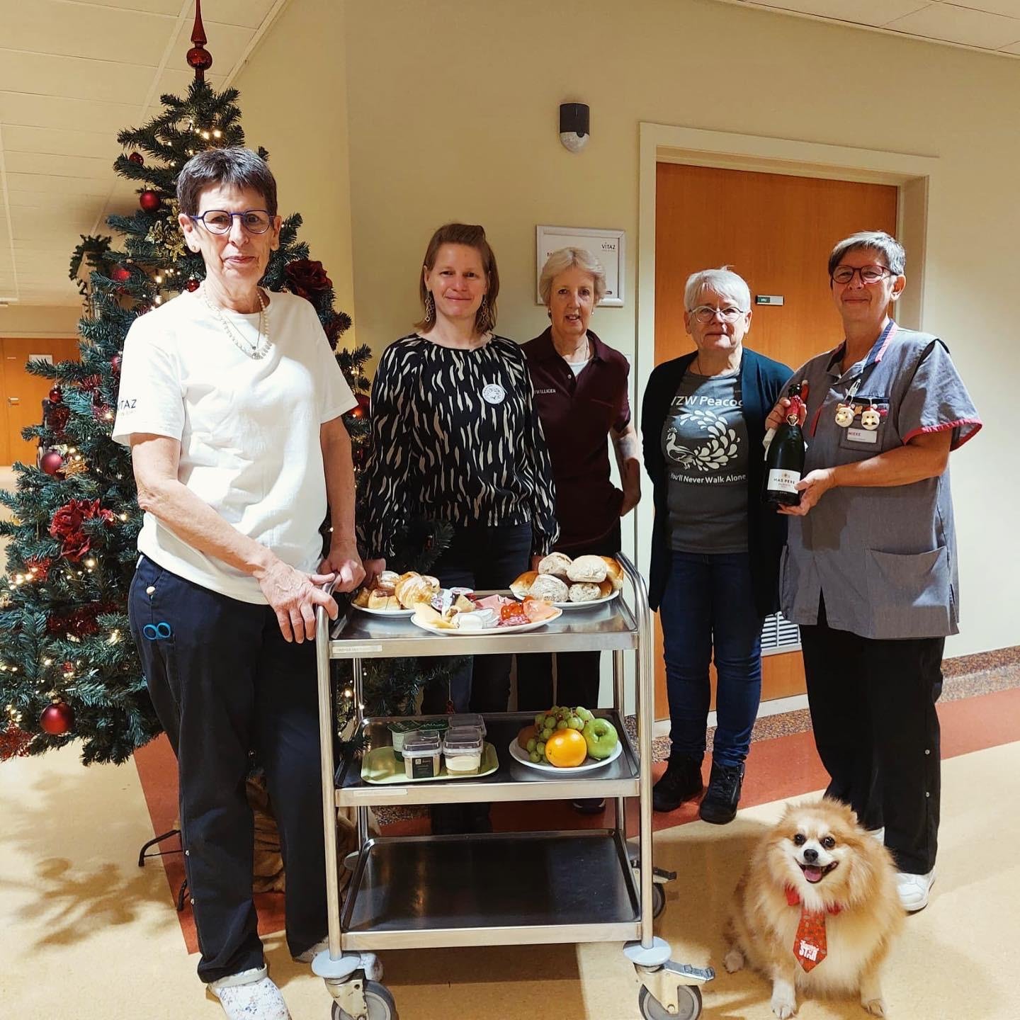 Vijf mensen met een kerstboom op de achtergrond, ze staan bij een serveerwagen met brood, fruit en kleine potjes, en houden een wijnfles vast. Er ligt een hond voor de serveerwagen.