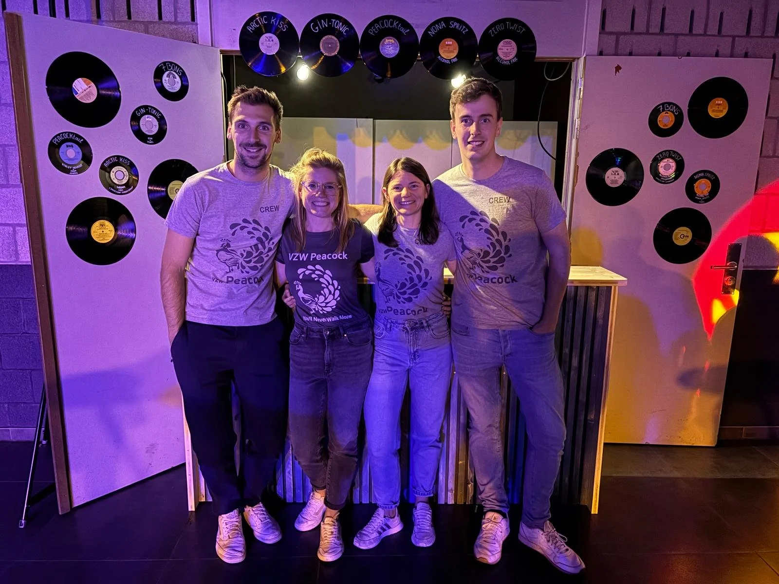 Vijf mensen poseerden in een groep, vier dragen grijze T-shirts met 'VZW Peacock' erop, de ene vrouw draagt een donkerblauwe T-shirt. Ze staan voor een achtergrond met platen van vinyl in een lichte, kleurrijke omgeving.