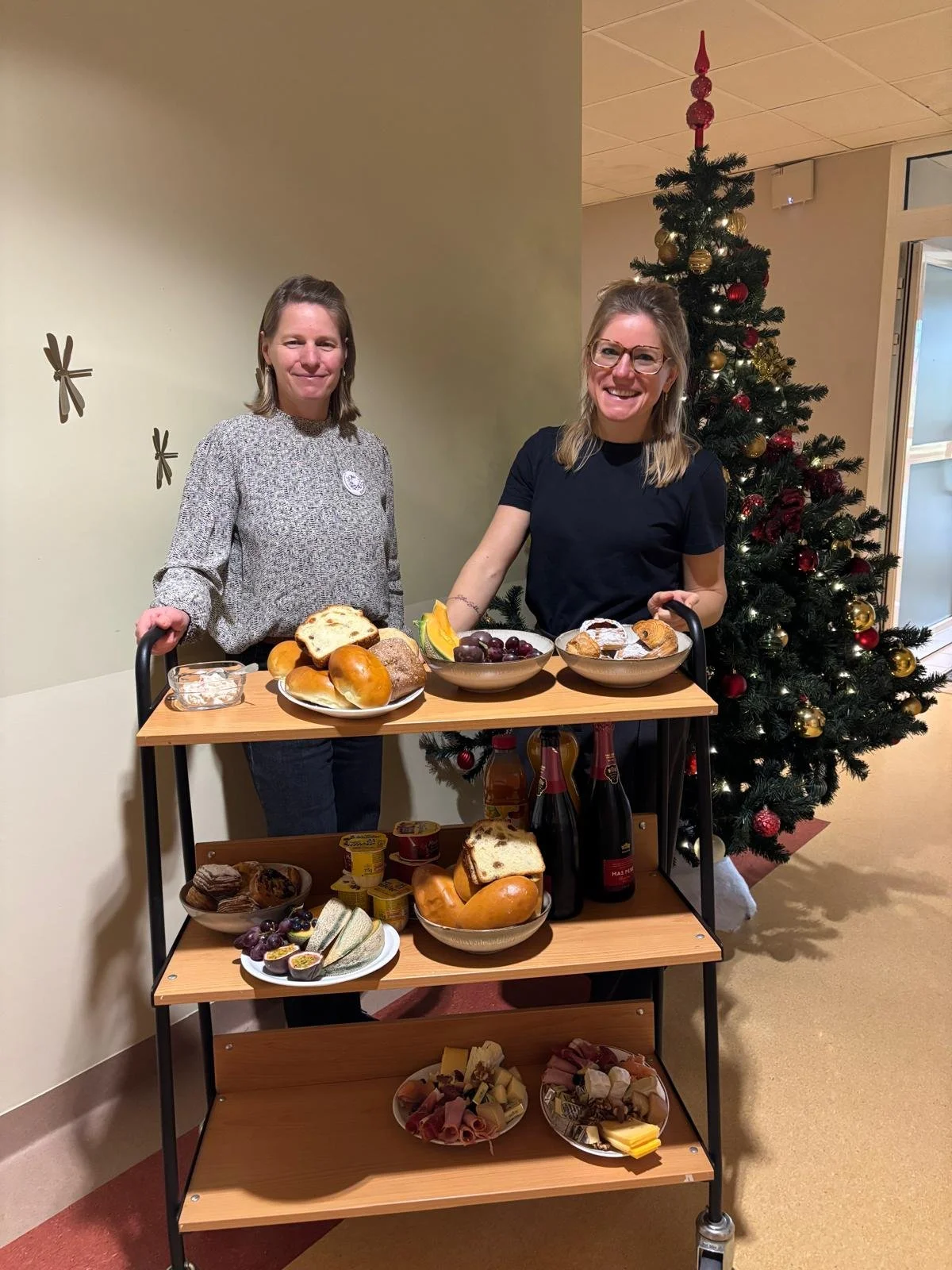 Twee vrouwen staan naast een kar met eten, met een versierde kerstboom op de achtergrond.