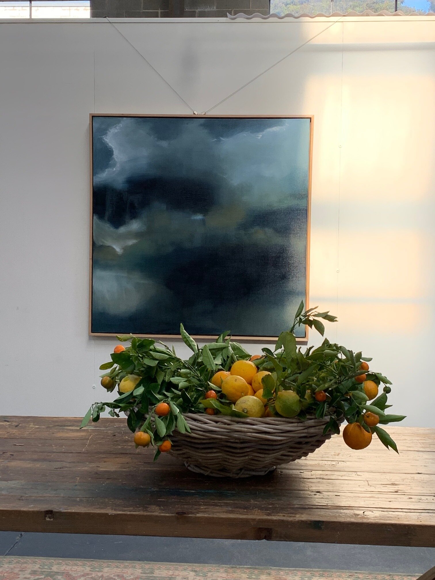 A basket filled with lemons and orange fruits on a wooden table, with an abstract blue painting on the wall behind.