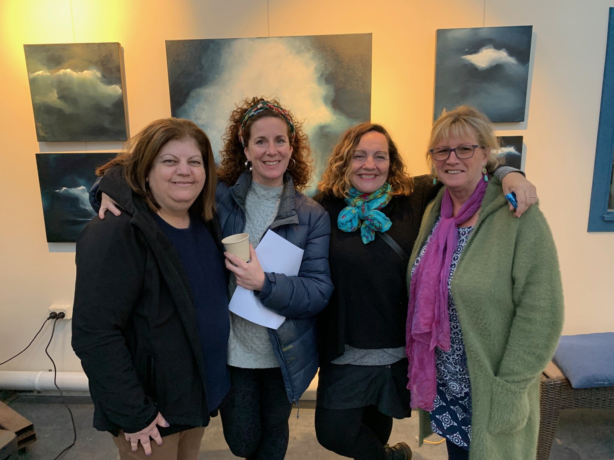 Four women standing together inside an art gallery, smiling, with abstract cloud paintings on the wall behind them.