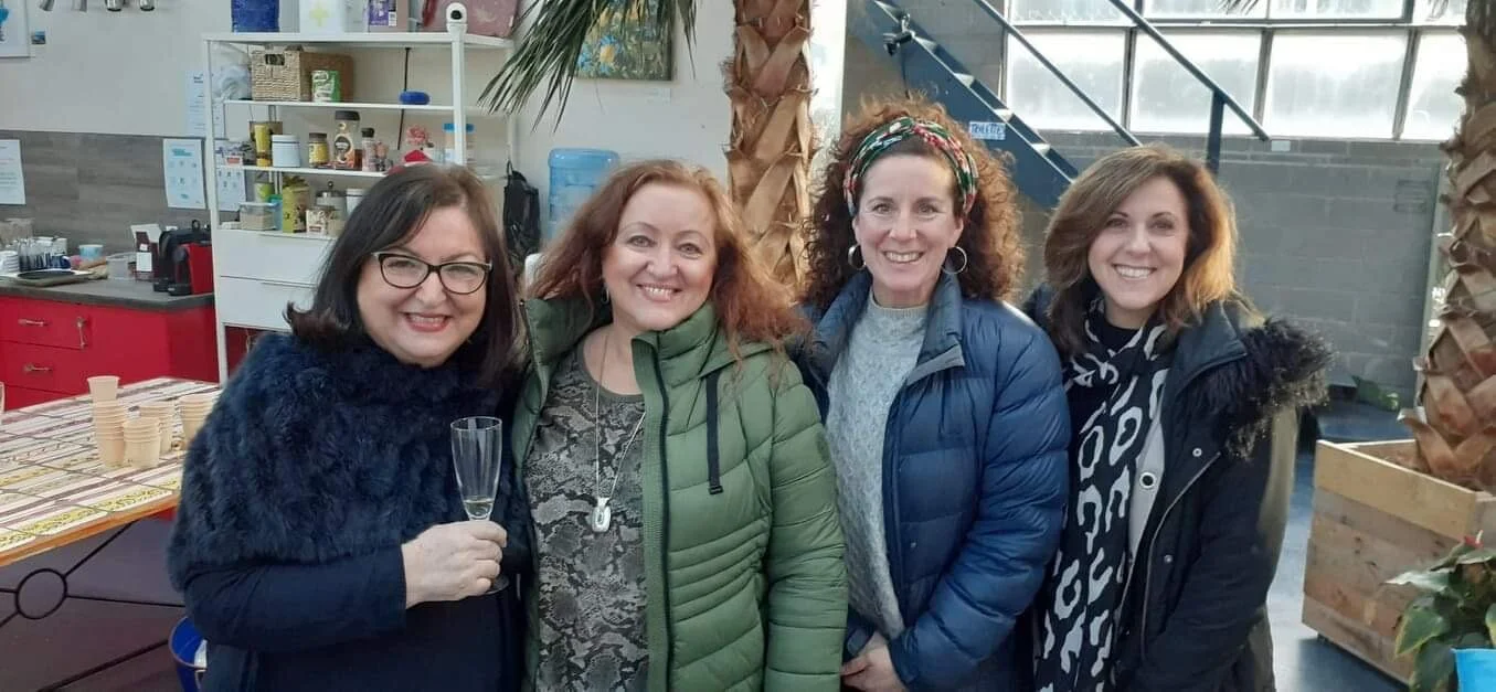 Four women smiling and standing together indoors, with one holding a champagne flute, in a room decorated with palm trees and a water dispenser.