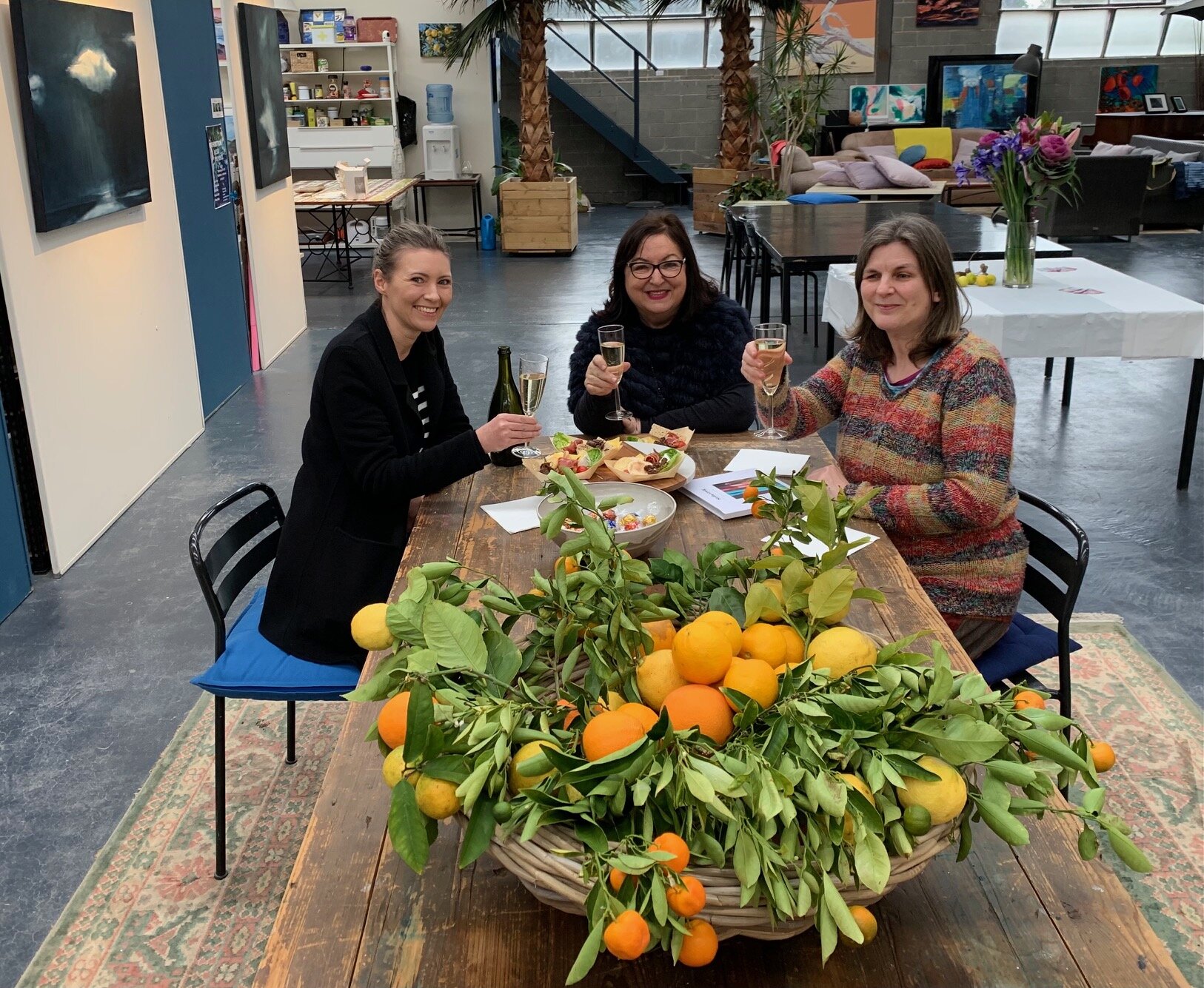 Three women sitting around a wooden table with a large basket of oranges and citrus fruits, smiling and holding glasses of champagne in an indoor space decorated with plants and artwork.