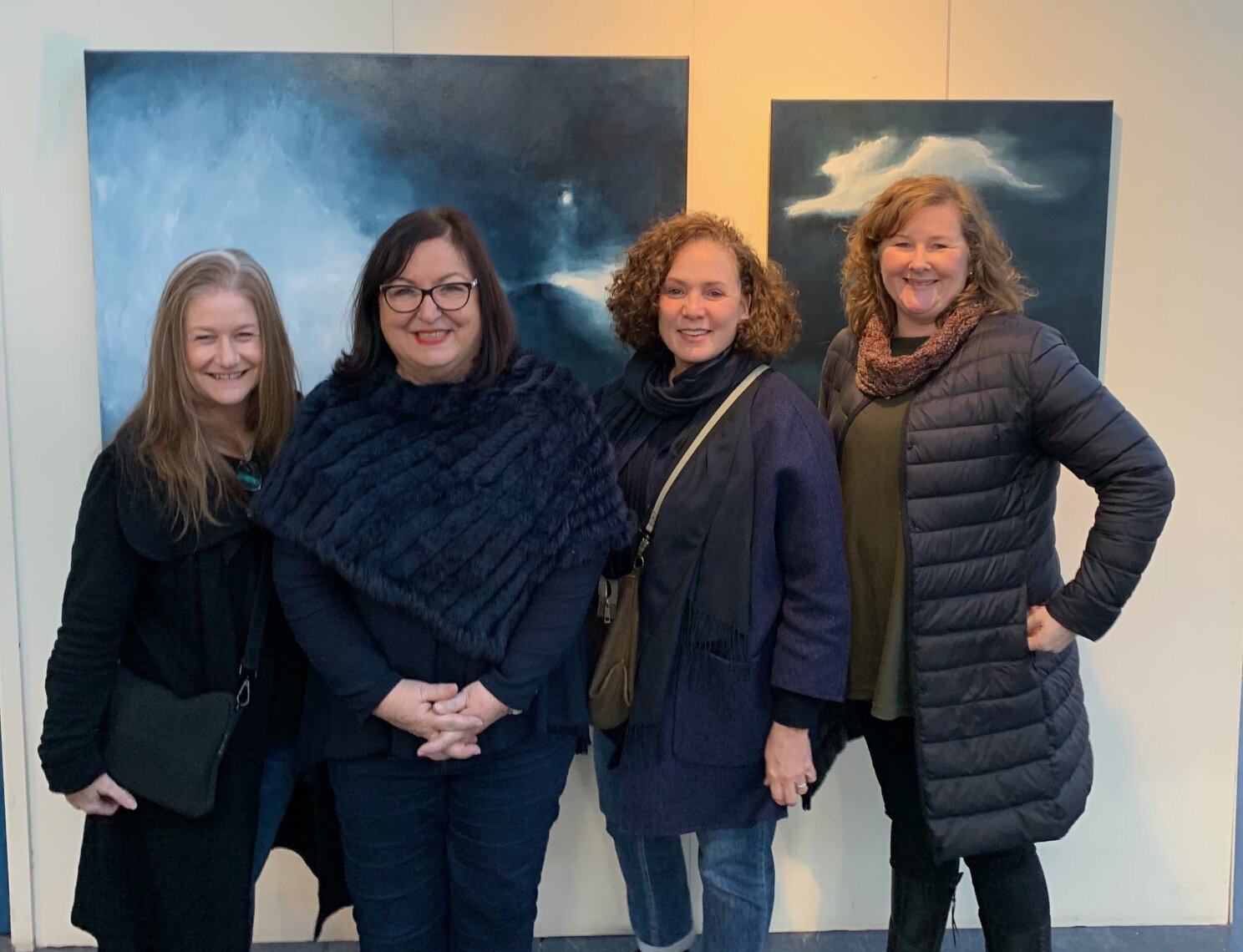 Four women standing together in front of two large abstract paintings at an art gallery, smiling at the camera.