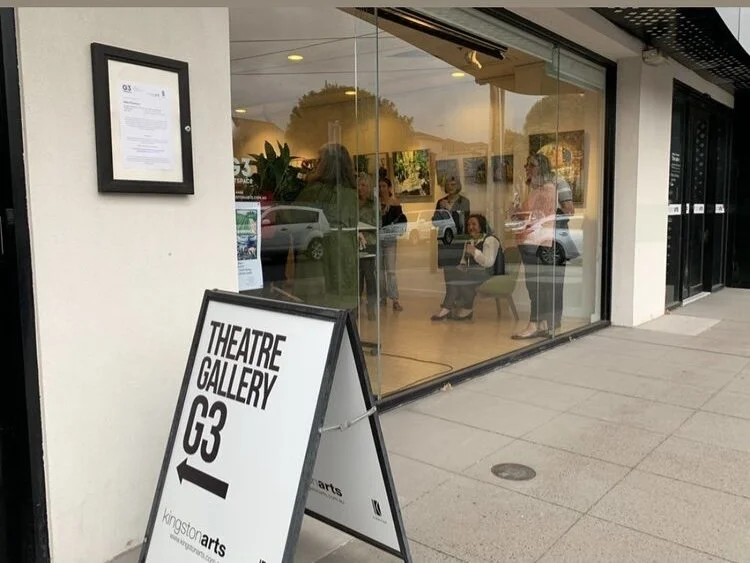 People sitting and standing inside an art gallery through large glass windows, with a sign outside that says 'Theatre Gallery G3' with an arrow pointing left, and another frame on the wall displaying a document.
