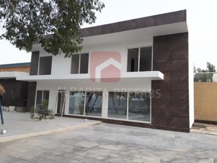 Modern two-story prefabricated house with large glass windows and white/brown exterior walls executed by Alrabita Prefab Co. بيت جاهز دورين مودرن مع شبابيك كبيرة وواجهة بلمسة خشبية تم تنفيذه من قبل مصنع الرابطة للبيوت الجاهزة