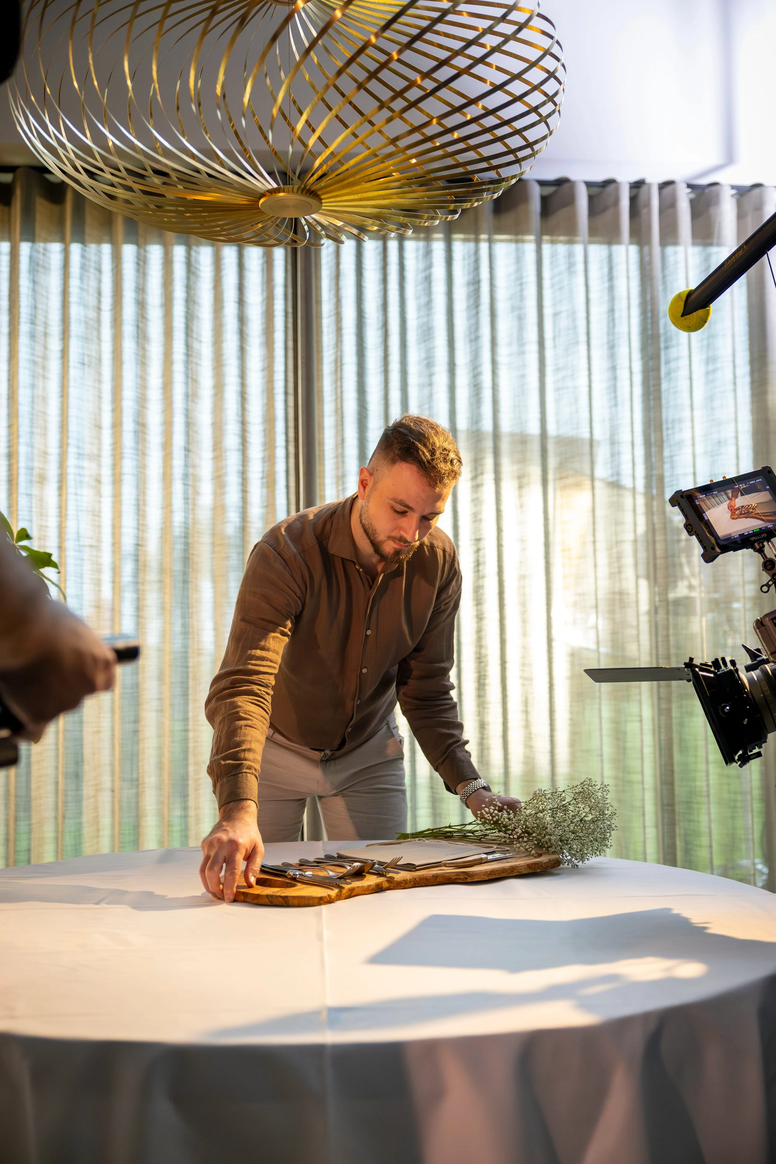 Man arranging cutlery, flowers, and a cutting board on a white-covered table in front of a camera and film crew, with curtains and a large ceiling light in the background.