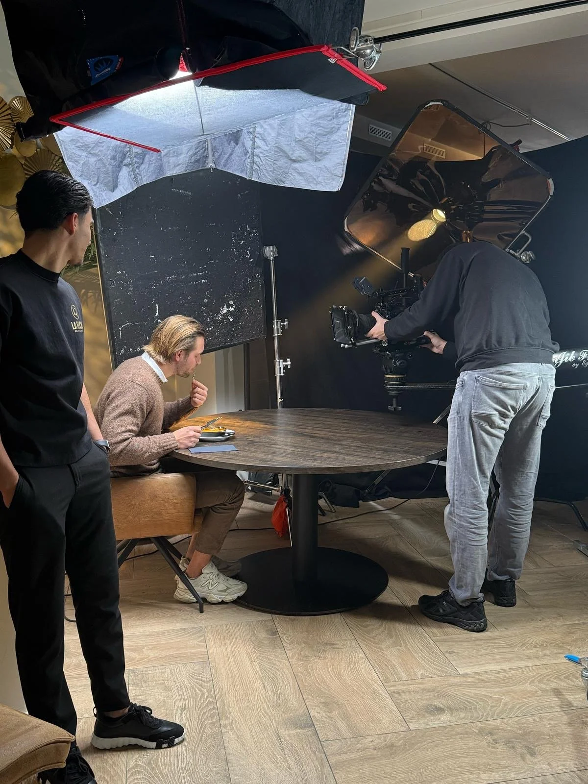 A man in a brown sweater is sitting at a round table, eating food, while two other men are standing, one holding a camera and the other observing, during an indoor video shoot setup with lighting equipment and large mirror reflectors.