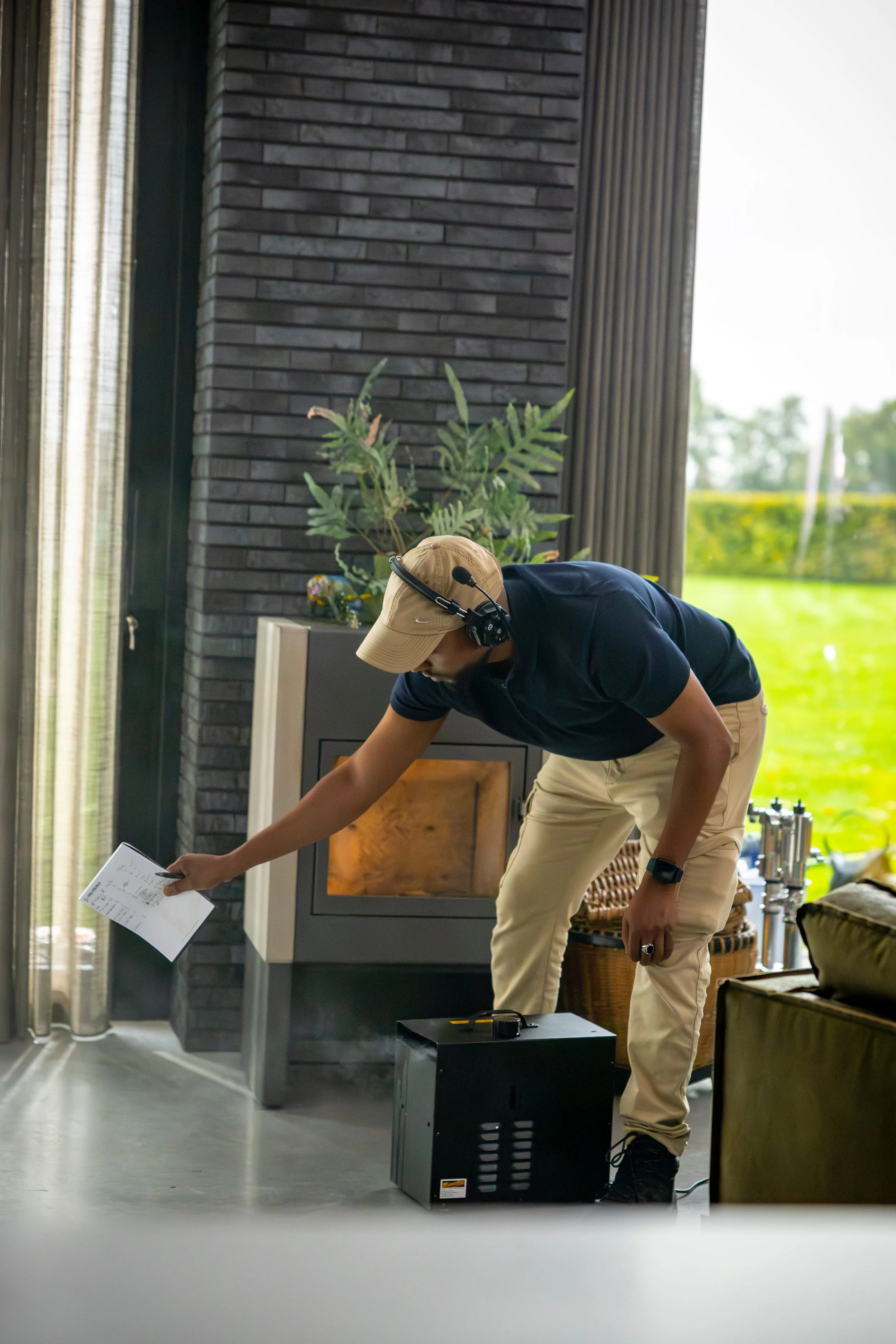 A person wearing a tan cap, navy polo shirt, and beige pants is adjusting a black portable device on the floor in a modern room with black brick walls, a fireplace, and large windows revealing green outdoor scenery. The person is wearing a headset an