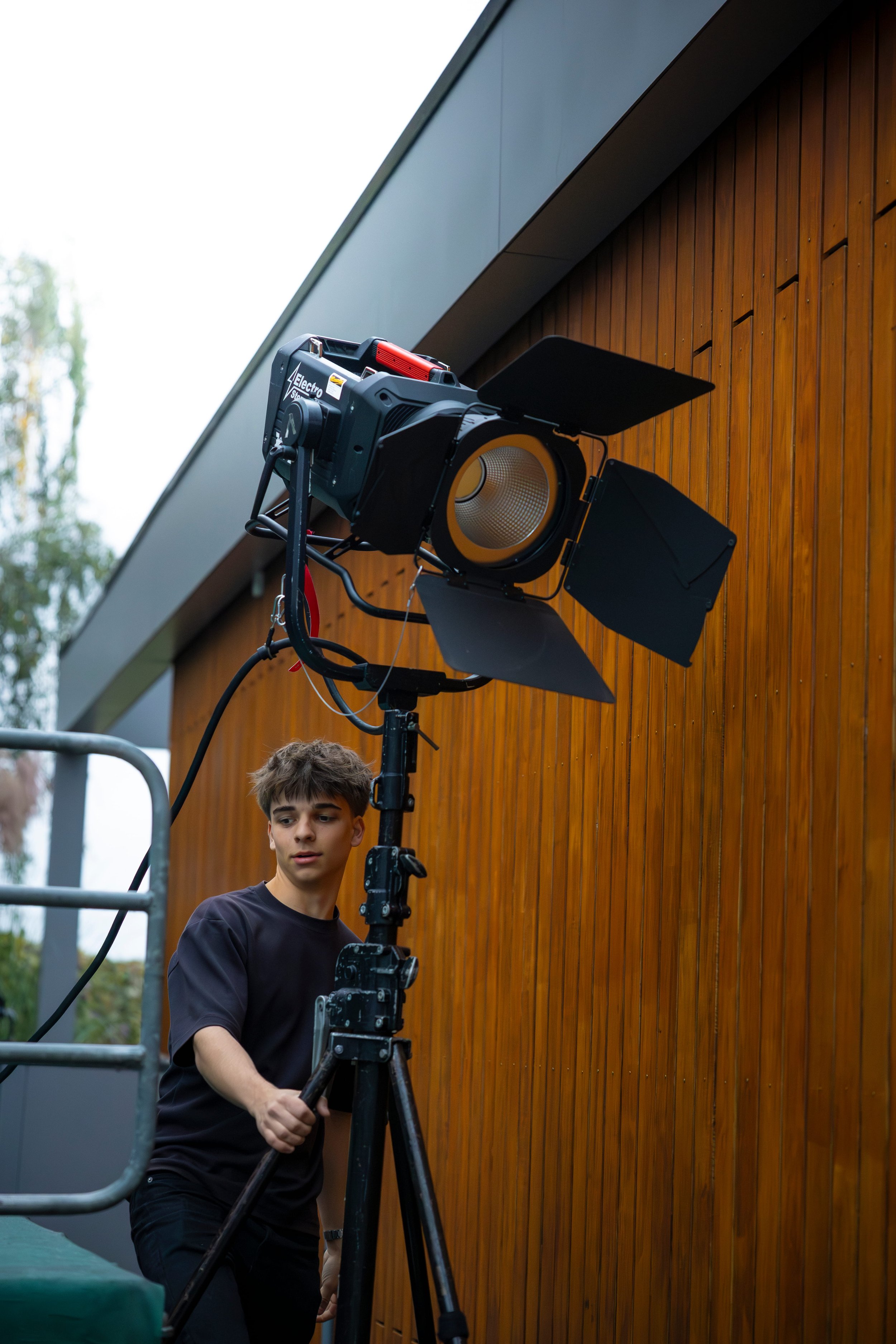 A young man adjusting a large professional studio light outside a building with wooden siding.