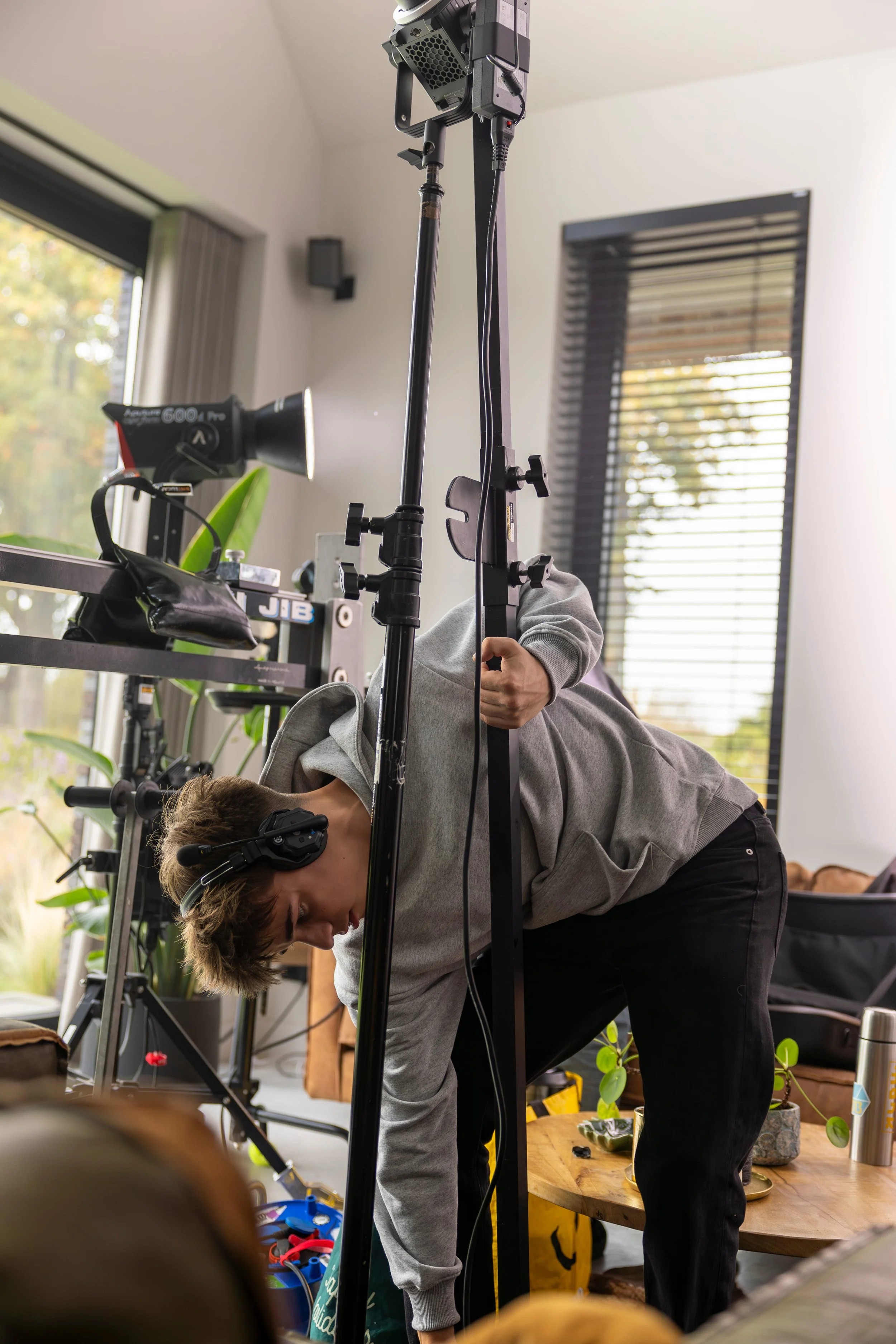A person in a gray hoodie, wearing headphones, is adjusting equipment or standing behind a camera on a tripod in a home office or studio with a large window and indoor plants.