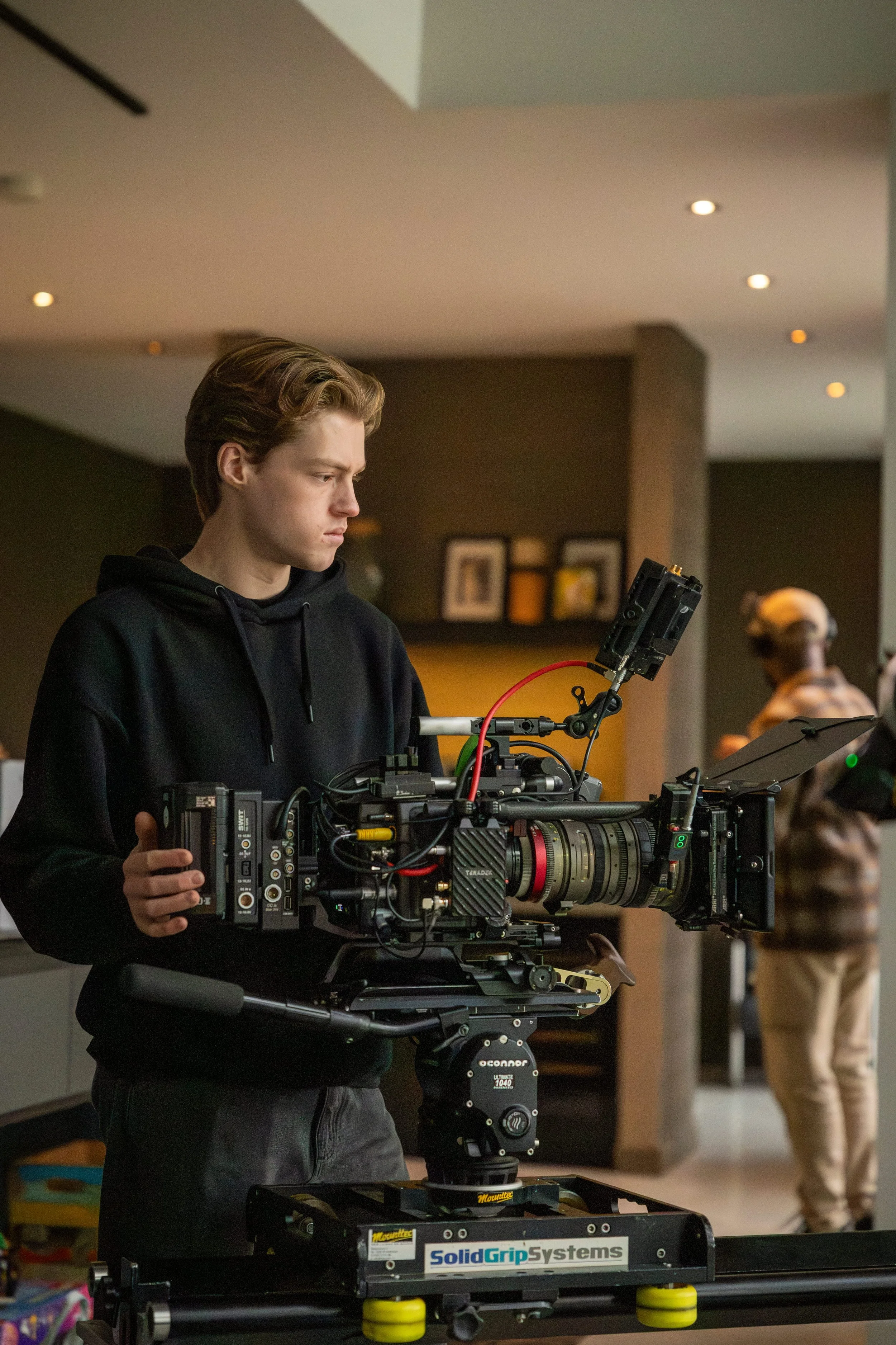A young man with light skin and brown hair operating a professional film camera indoors, with other people blurred in the background.