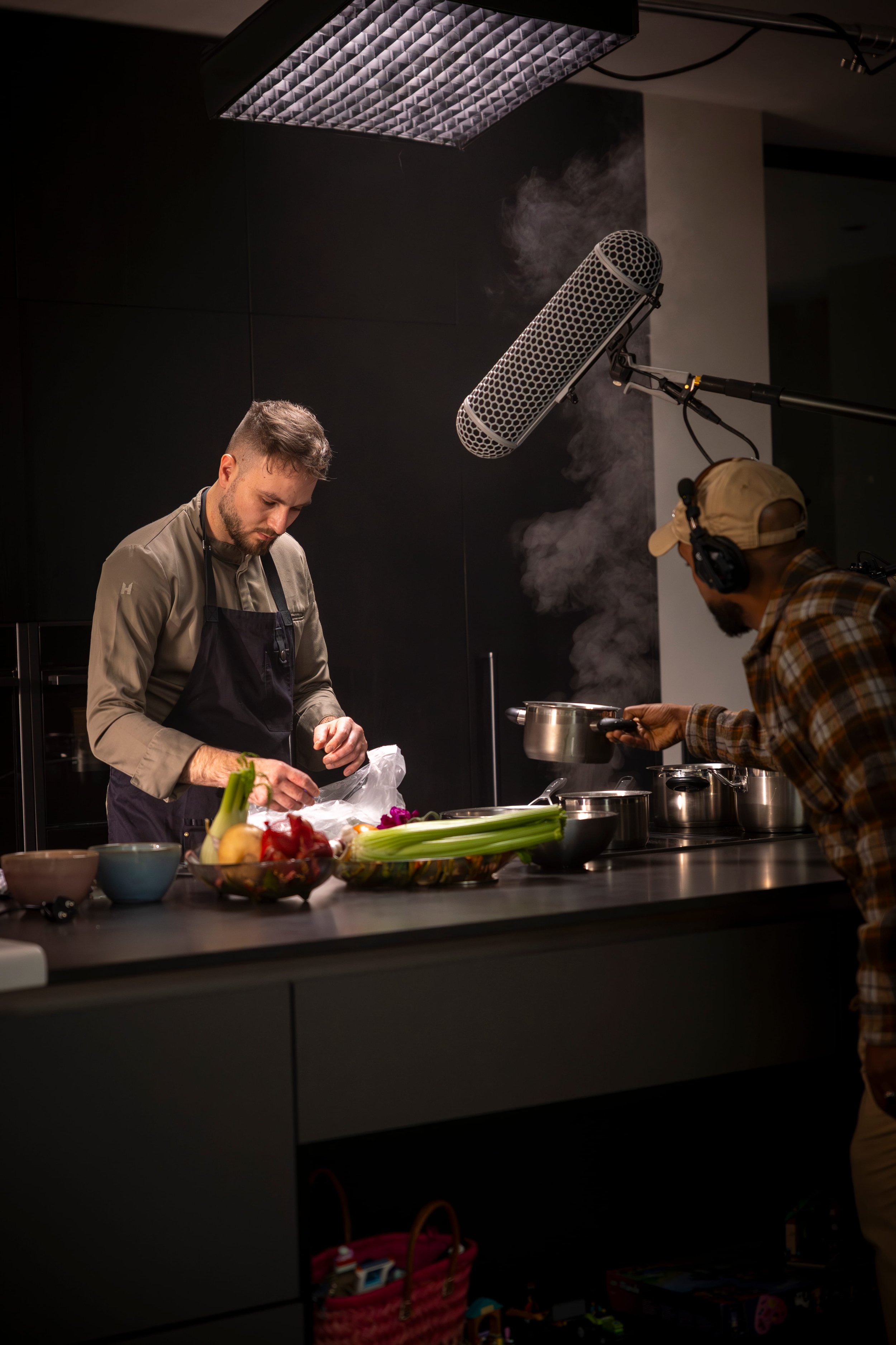 Two men working in a professional kitchen with a black background, preparing food at a countertop with vegetables and bowls, one man is stirring a pot while the other is adding ingredients, and a large microphone is above them for filming.